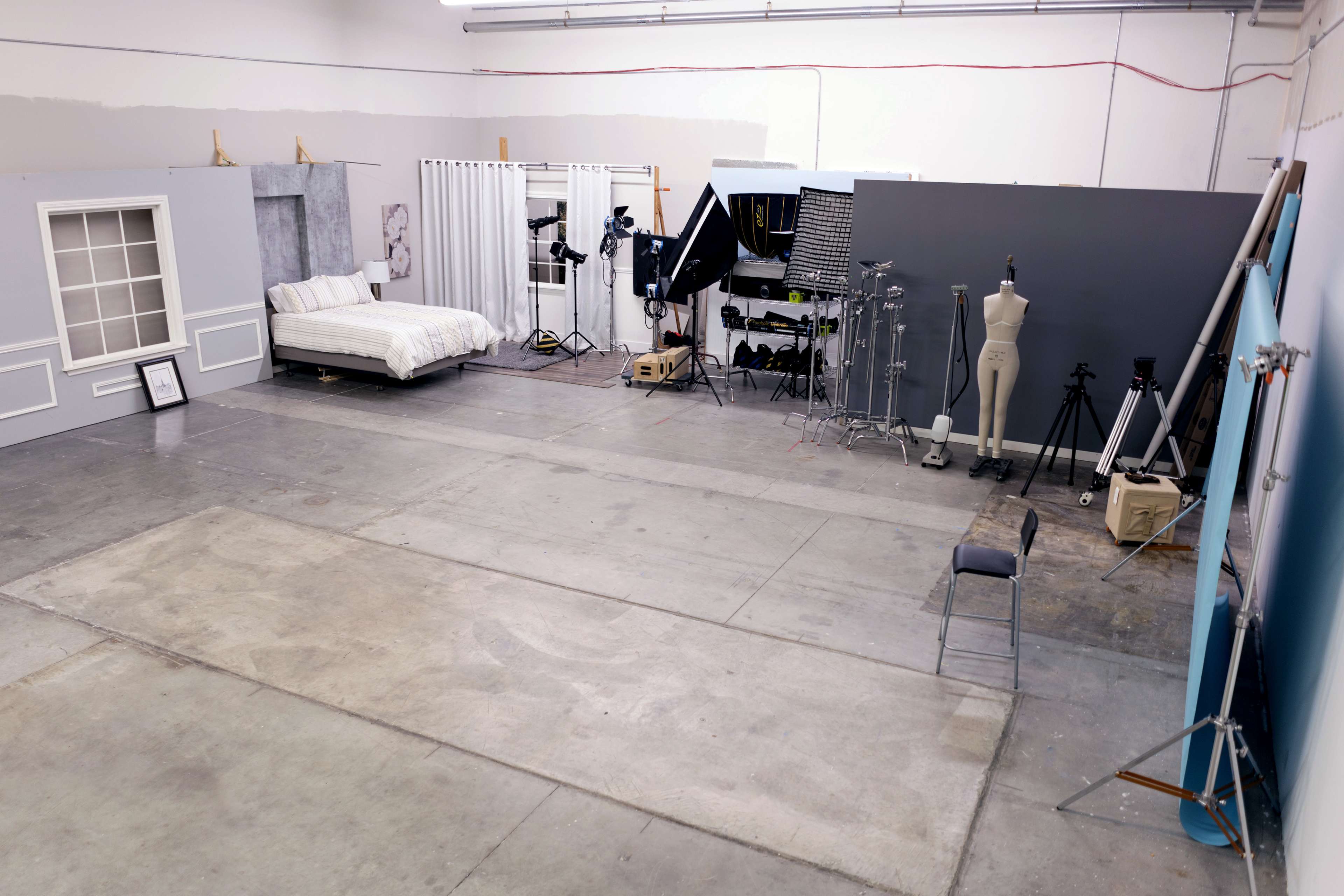 A spacious photography studio featuring a bed, various lighting equipment, a clothing mannequin, and several tripods arranged against plain gray walls.