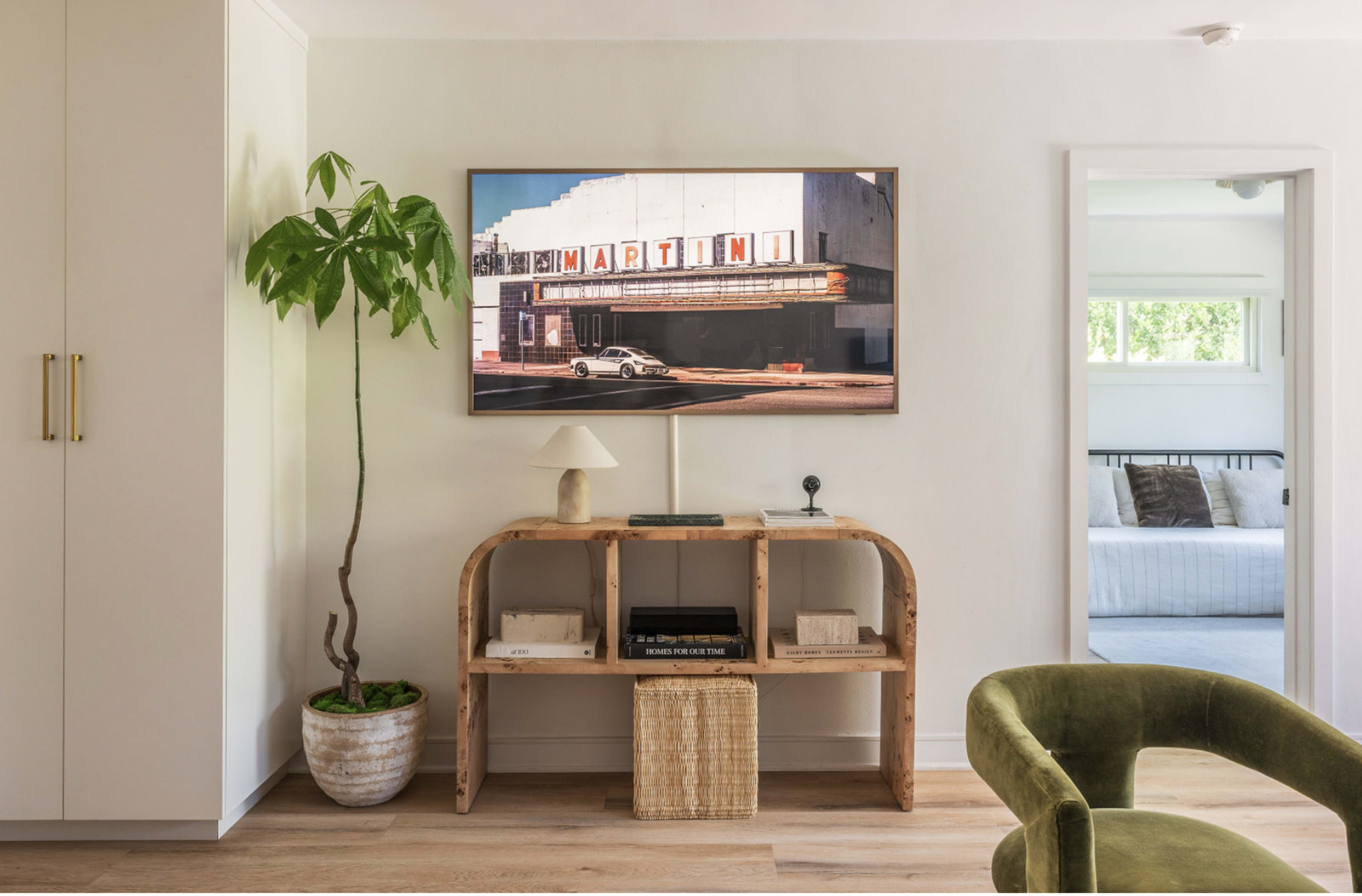 A modern living room features a TV mounted above a wooden console table, with a plant beside it and a doorway leading to another room in the background.