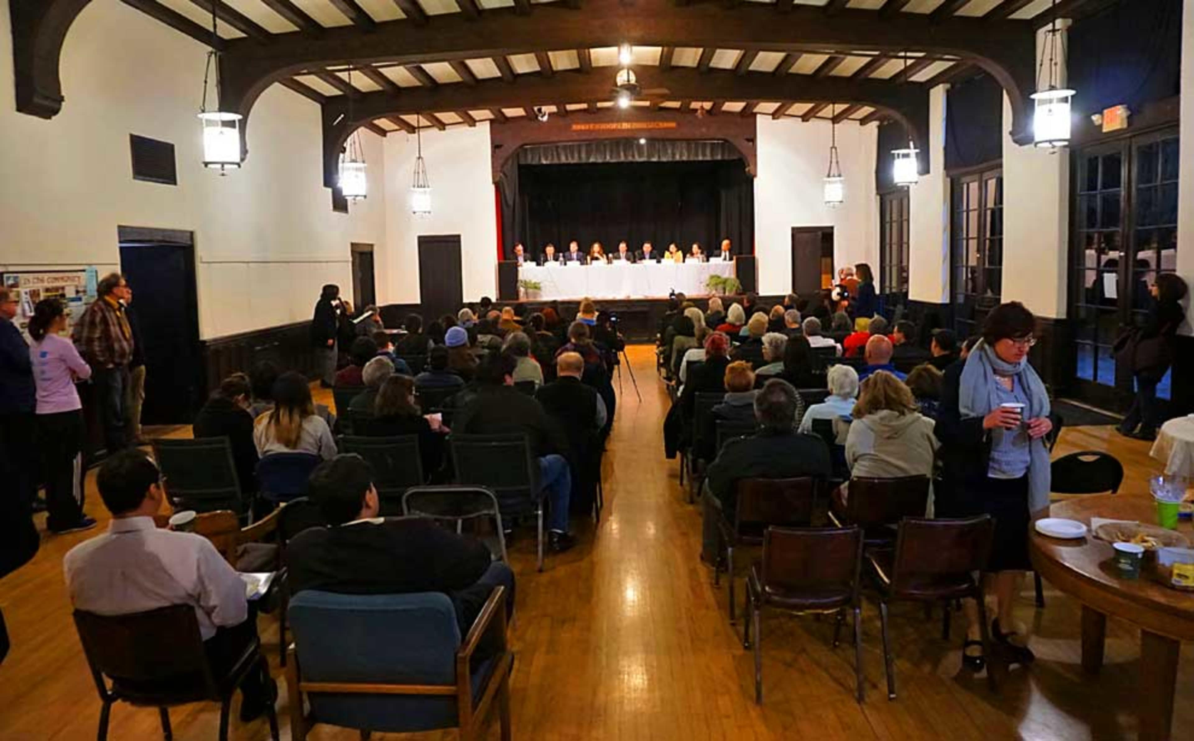 A large group of people sits in an auditorium facing a panel of speakers at the front.
