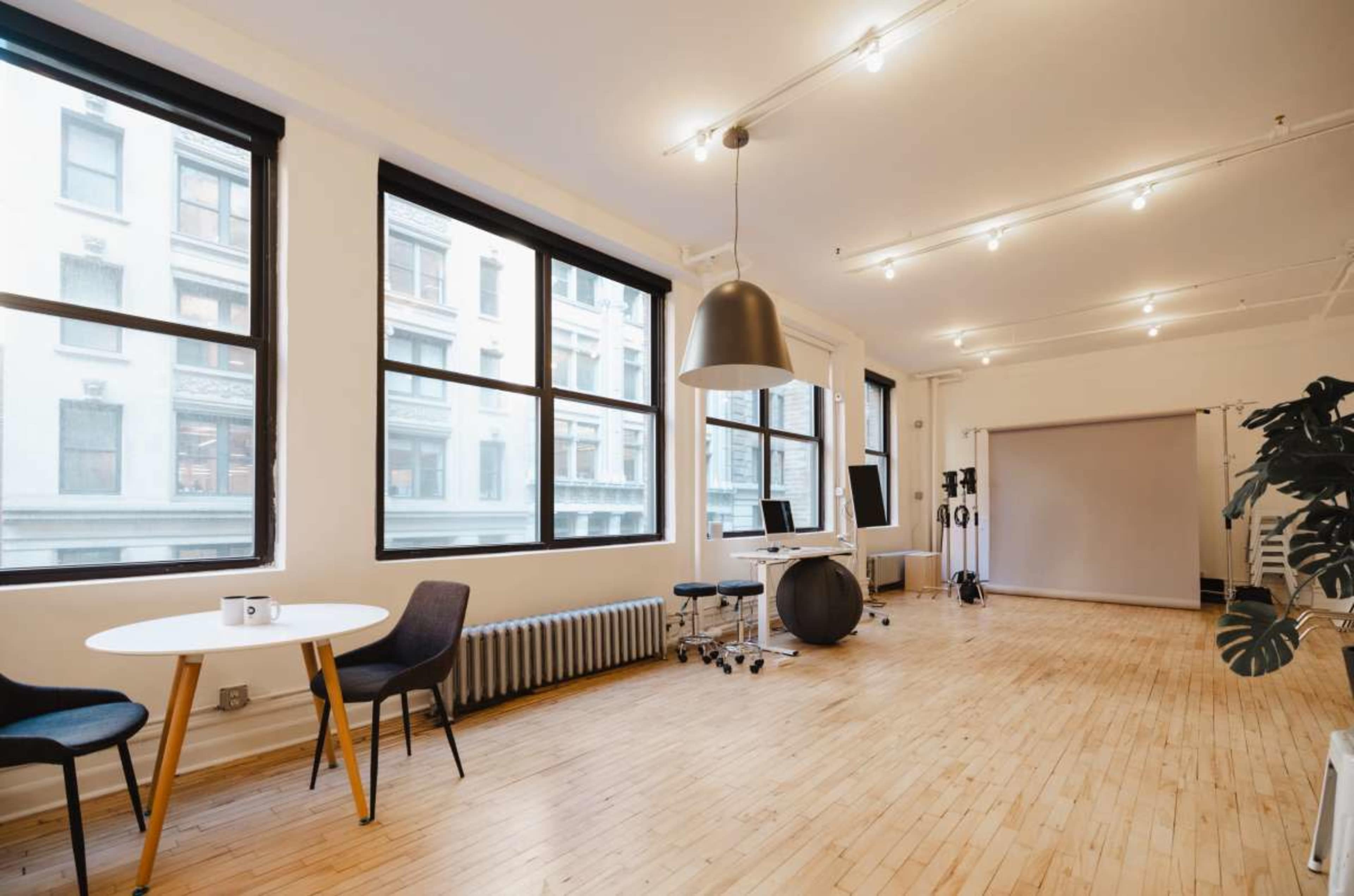 The image shows a well-lit, minimalistic studio space with large windows, a round table, two chairs, a radiator, and a backdrop.