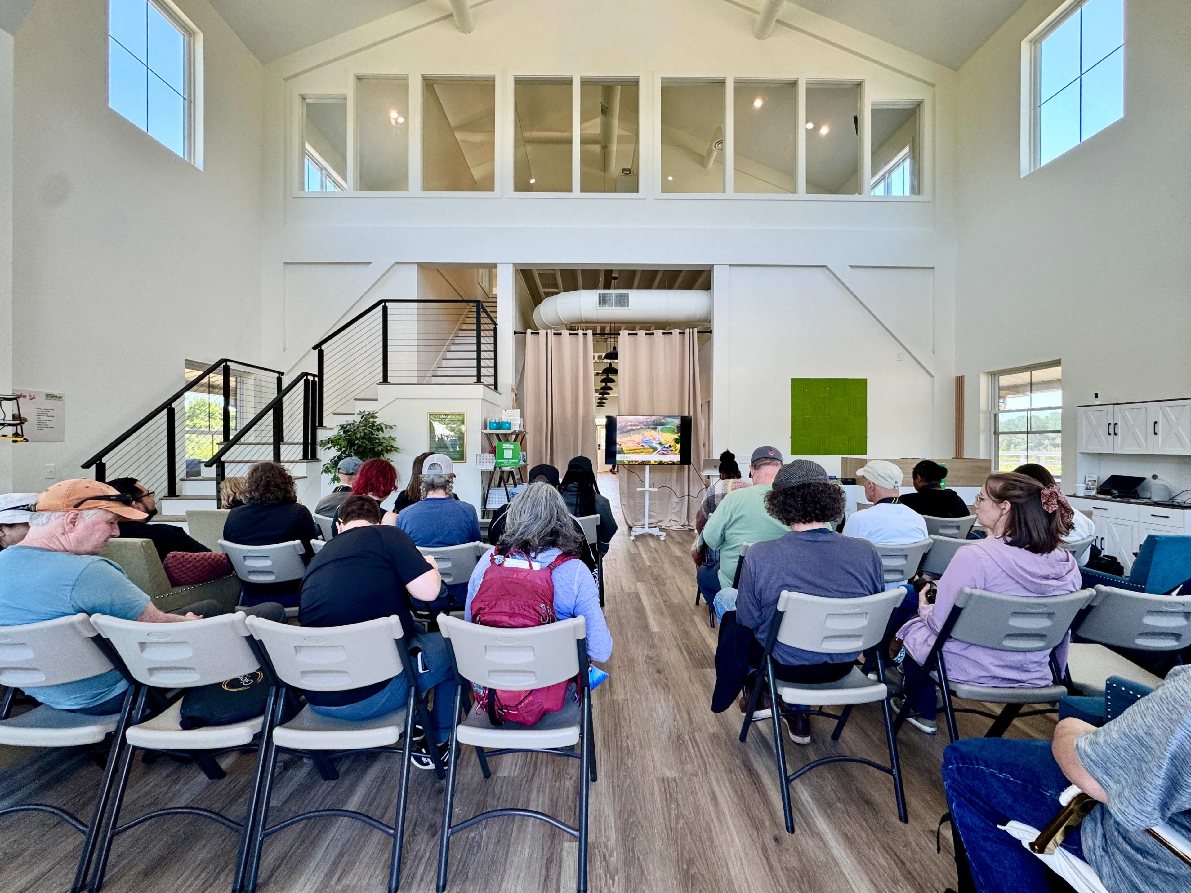 A group of people is seated in rows facing a large screen in a spacious, brightly lit indoor setting with high ceilings and large windows.