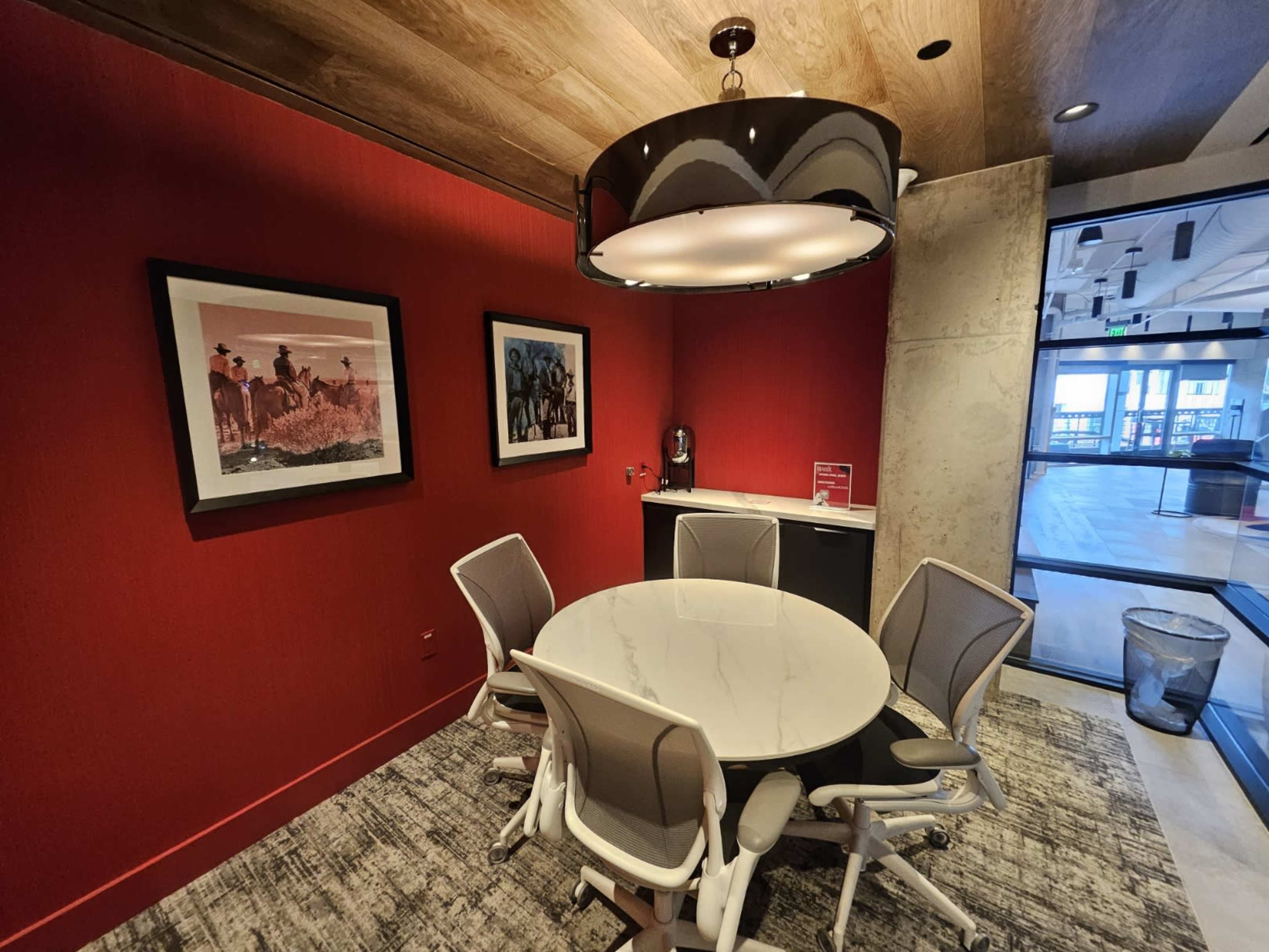 The image shows a modern conference room featuring a round white table surrounded by six chairs, with red walls and two framed pictures.