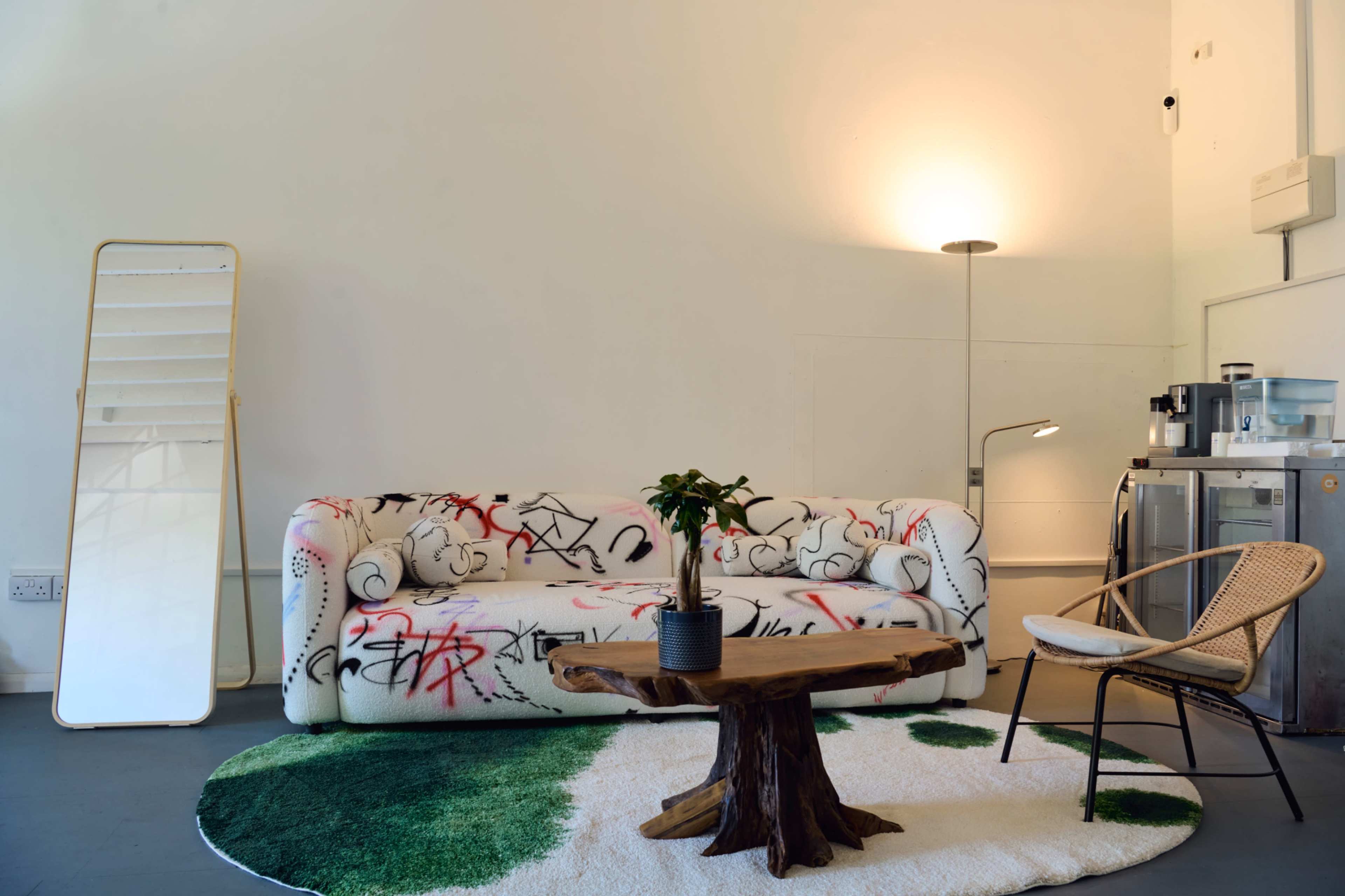 The image shows a modern interior with a graffiti-patterned couch, a round wooden coffee table, a potted plant, a floor lamp, and a glass display case against a plain wall.