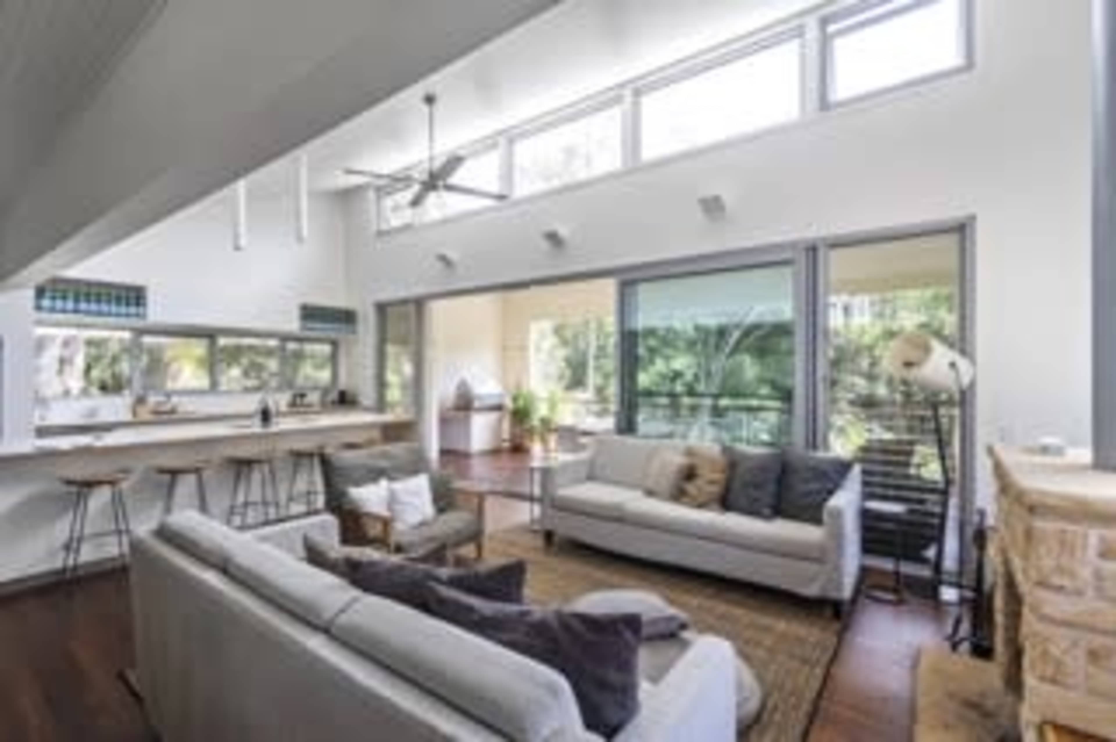 A bright, open-plan living room with two sofas, a coffee table, and large windows overlooking a green outdoor area.