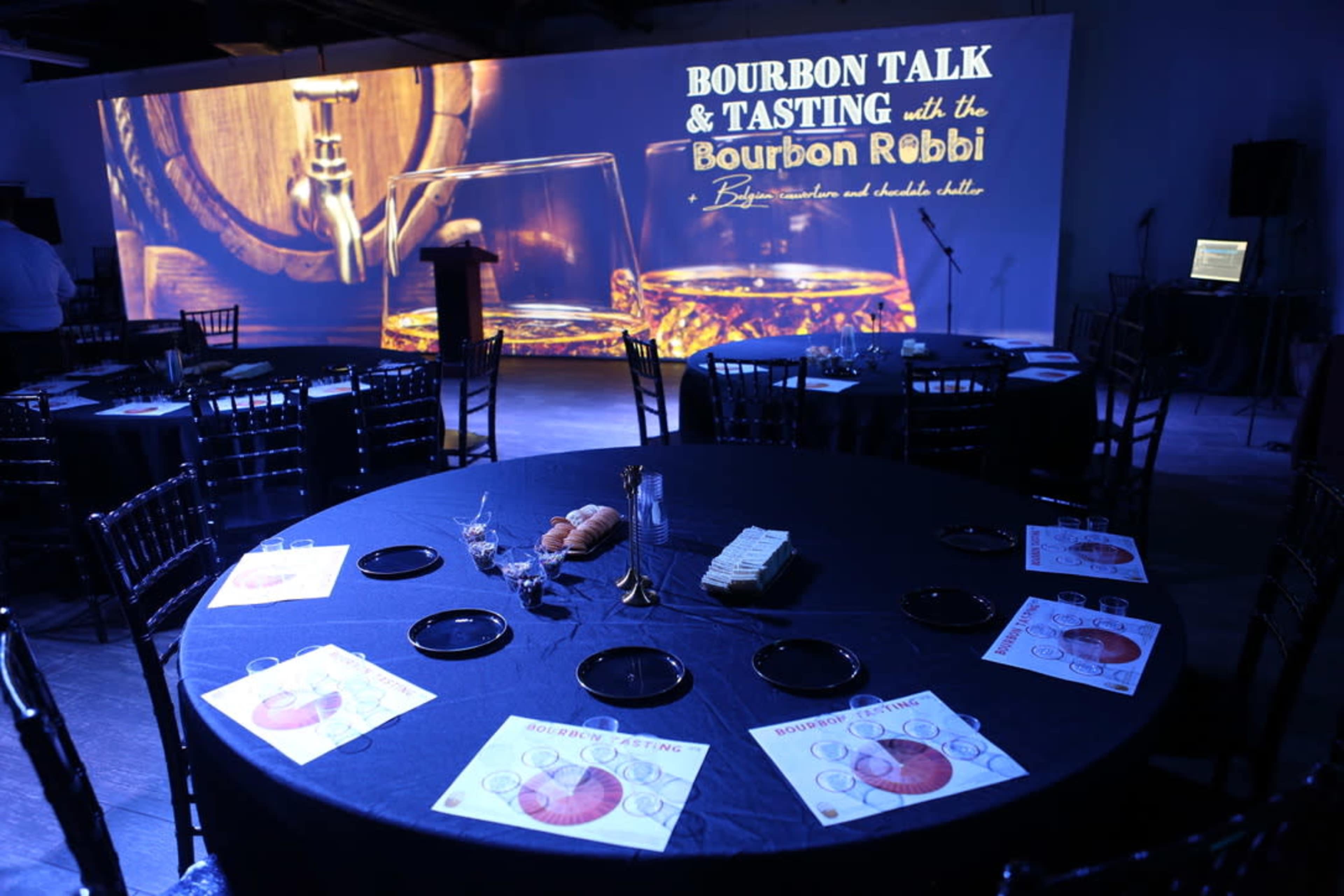 A round table is set with tasting materials and snacks in a dimly lit room featuring a large screen displaying "Bourbon Talk & Tasting."