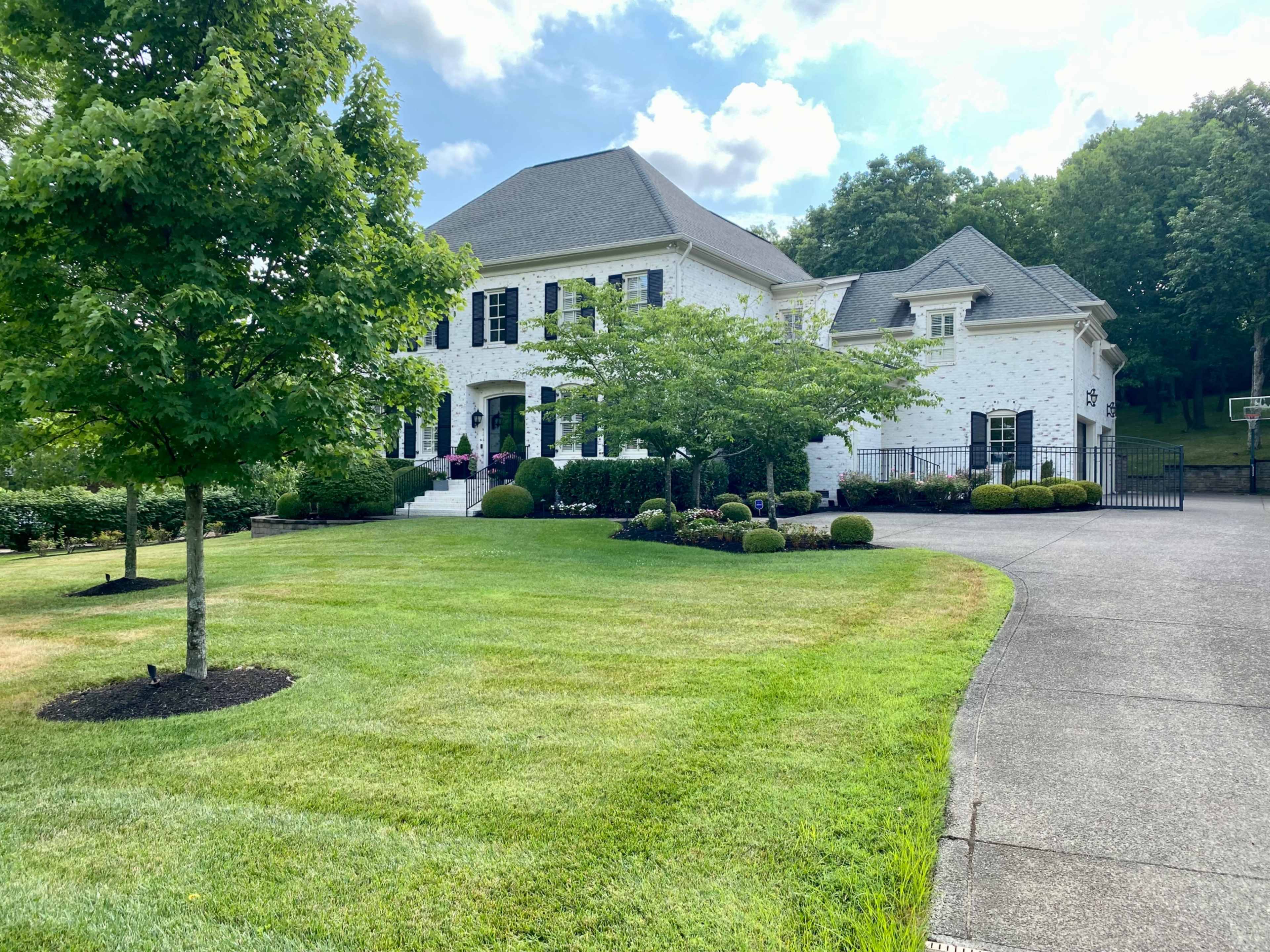 A large white house with black shutters and a manicured lawn, surrounded by trees and shrubbery.