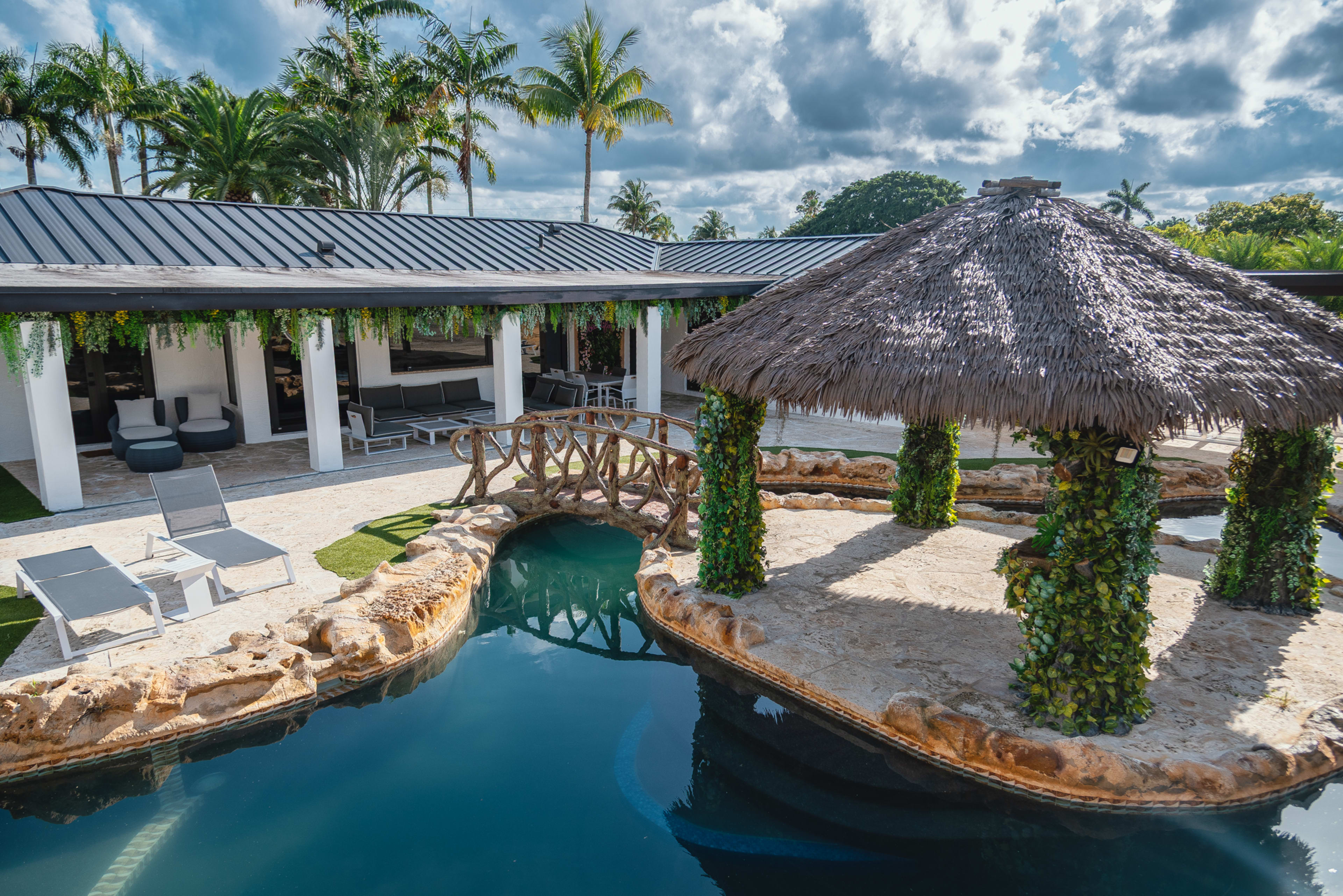 The image shows a modern outdoor pool area featuring a thatched gazebo, lounge chairs, and tropical landscaping.