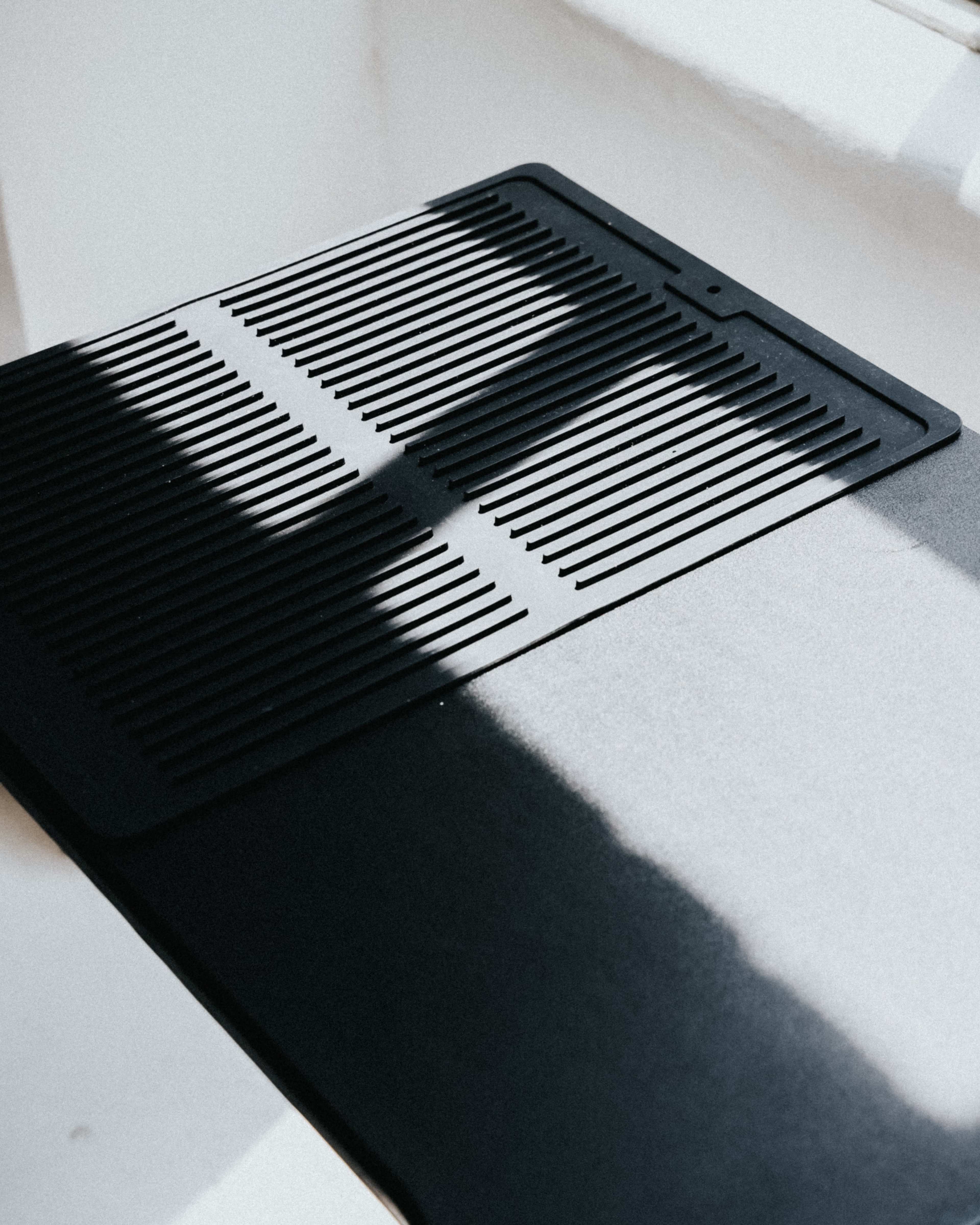 The image shows a black vent cover placed on a flat surface, with shadows cast across it.