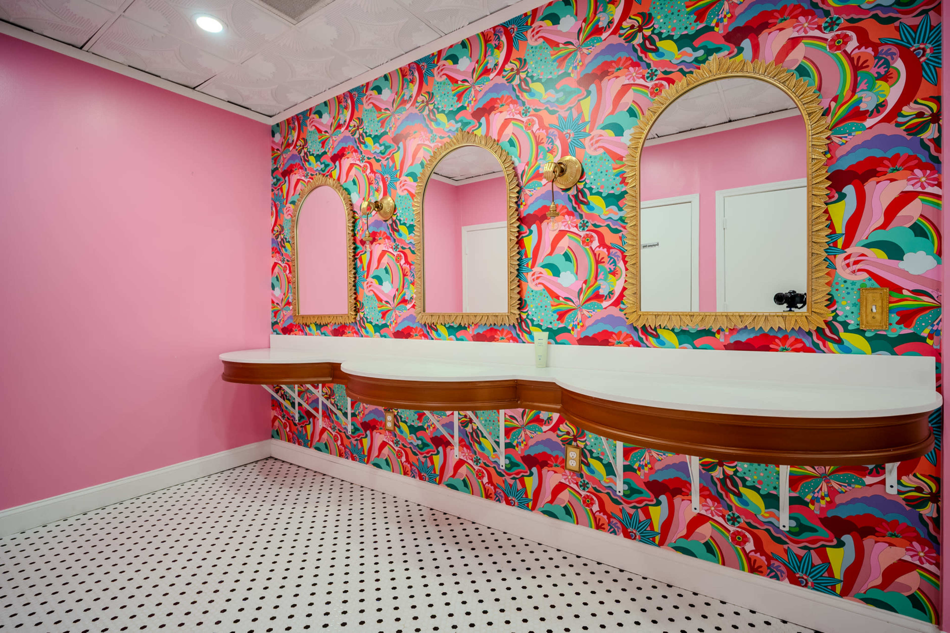 The image shows a vibrant restroom featuring a colorful, patterned wallpaper, three arched mirrors, and white sink countertops on wooden vanities.