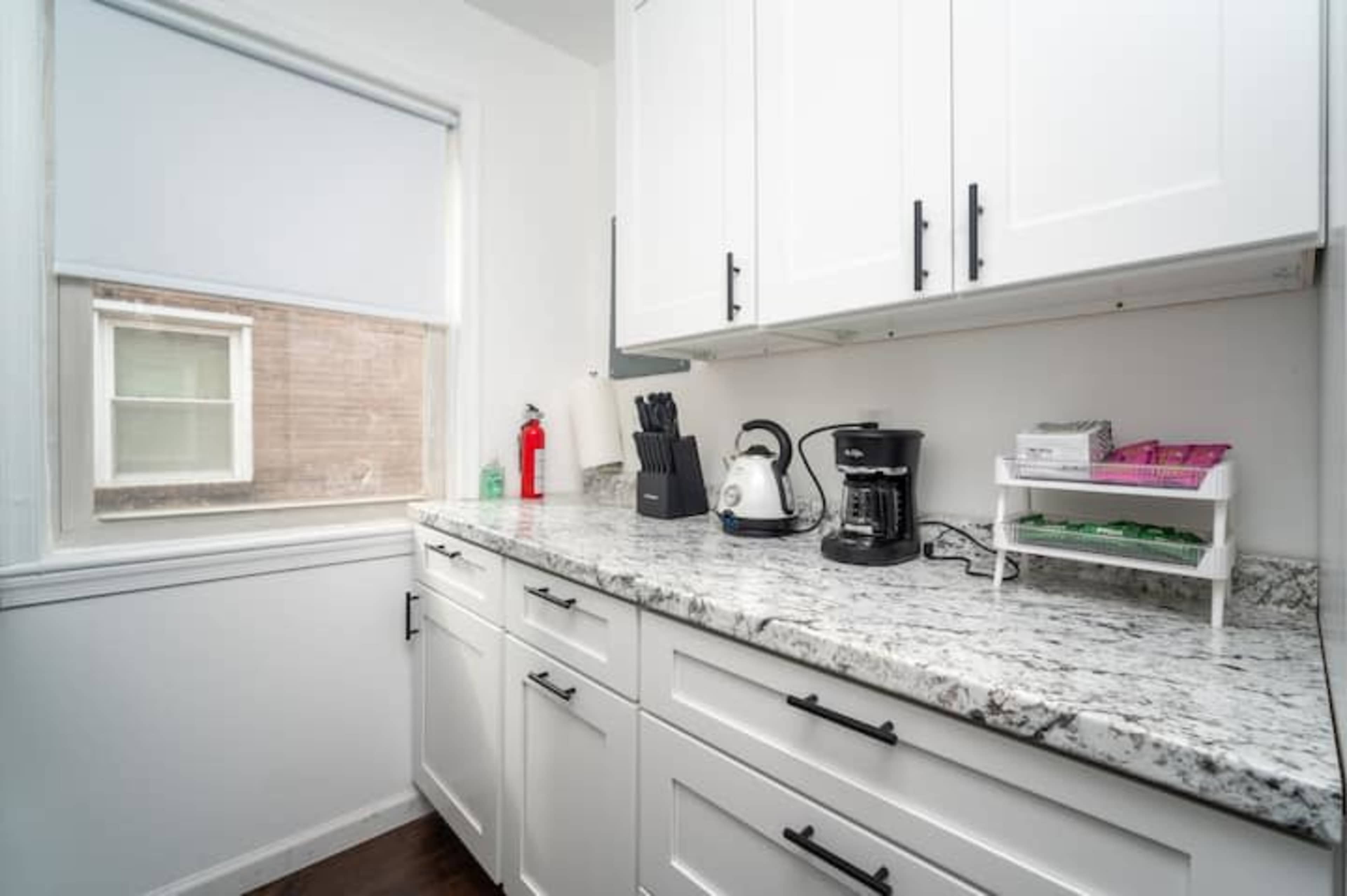 A small kitchen features white cabinets, a granite countertop, and appliances including a kettle and coffee maker, with a window providing natural light.