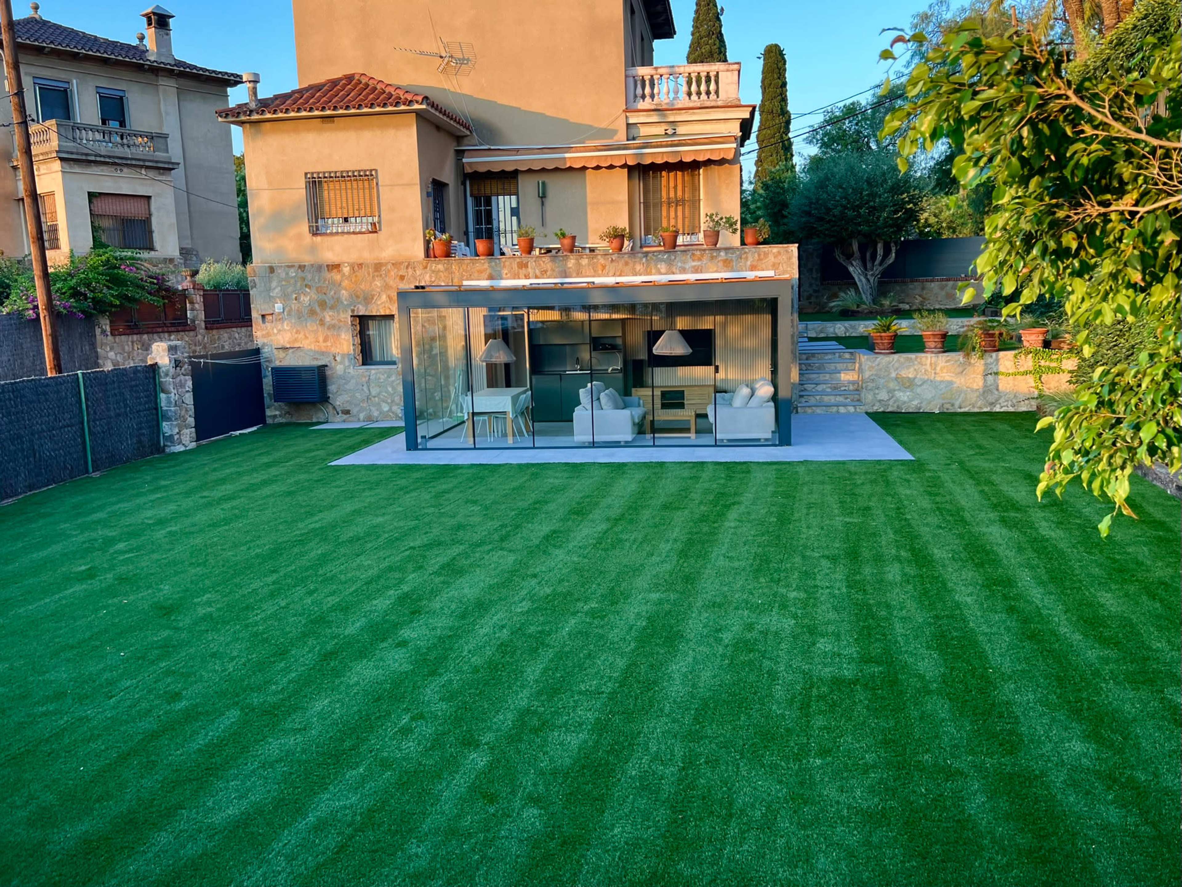 The image shows a modern glass structure surrounded by a manicured green lawn and a two-story house in the background.
