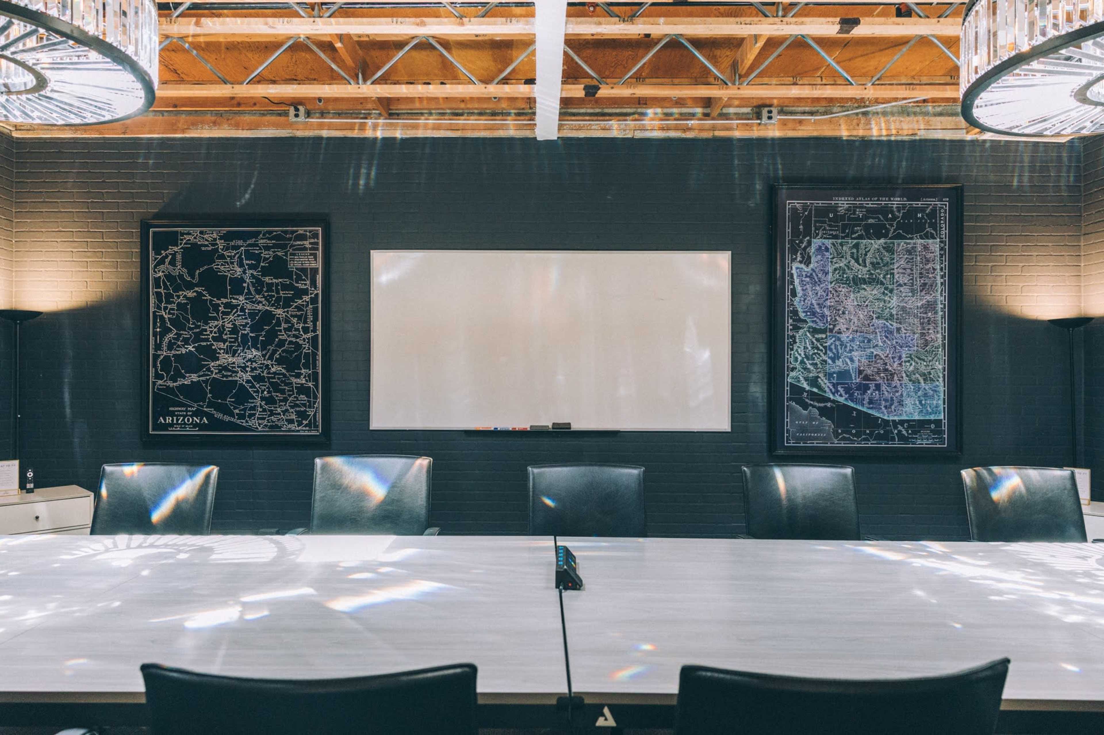 A modern conference room features a long table surrounded by black chairs, with two framed maps on the walls and a whiteboard in the center.
