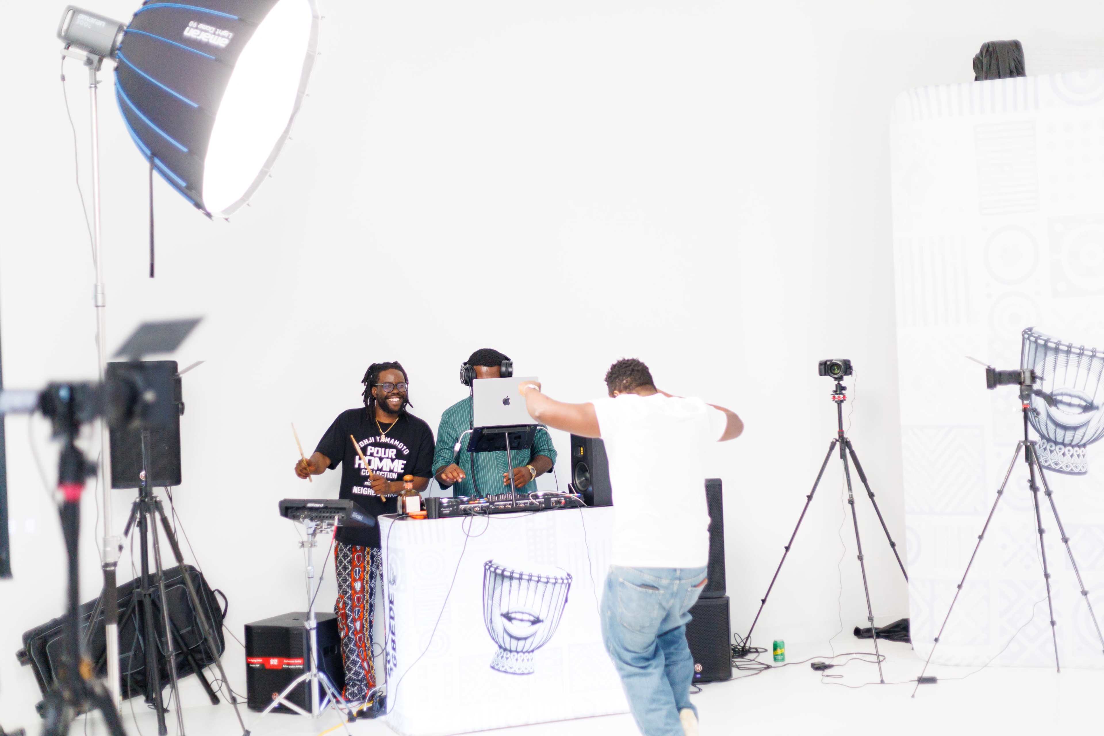 A DJ and a percussionist perform onstage while a person dances in front of them in a studio with a white backdrop.