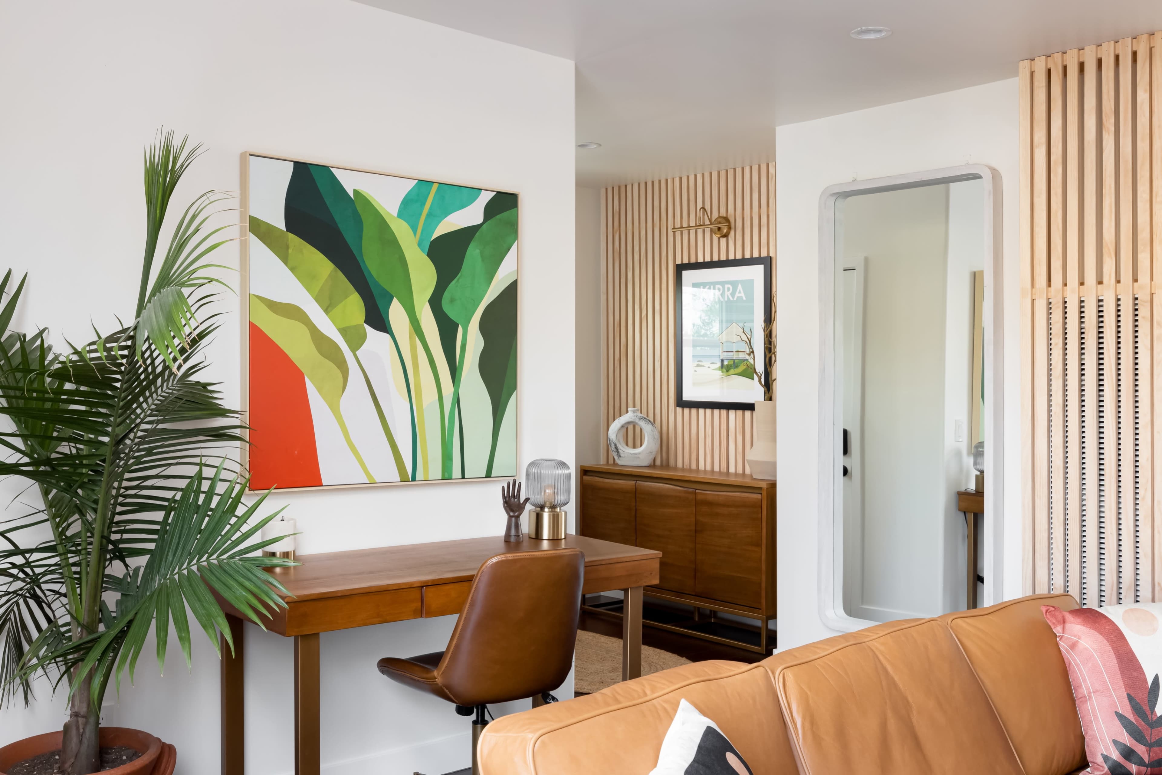 A modern living space features a desk with a brown chair, a large green and orange artwork on the wall, and a mirrored door leading to another room.