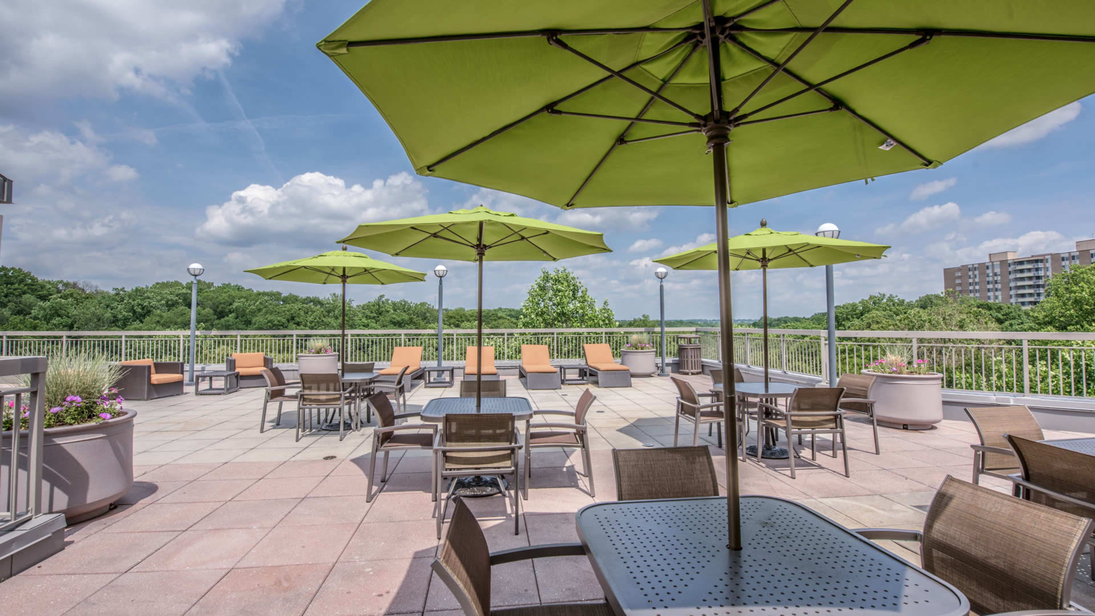 A rooftop terrace features several green umbrellas over tables and chairs, with a view of trees and buildings in the background.
