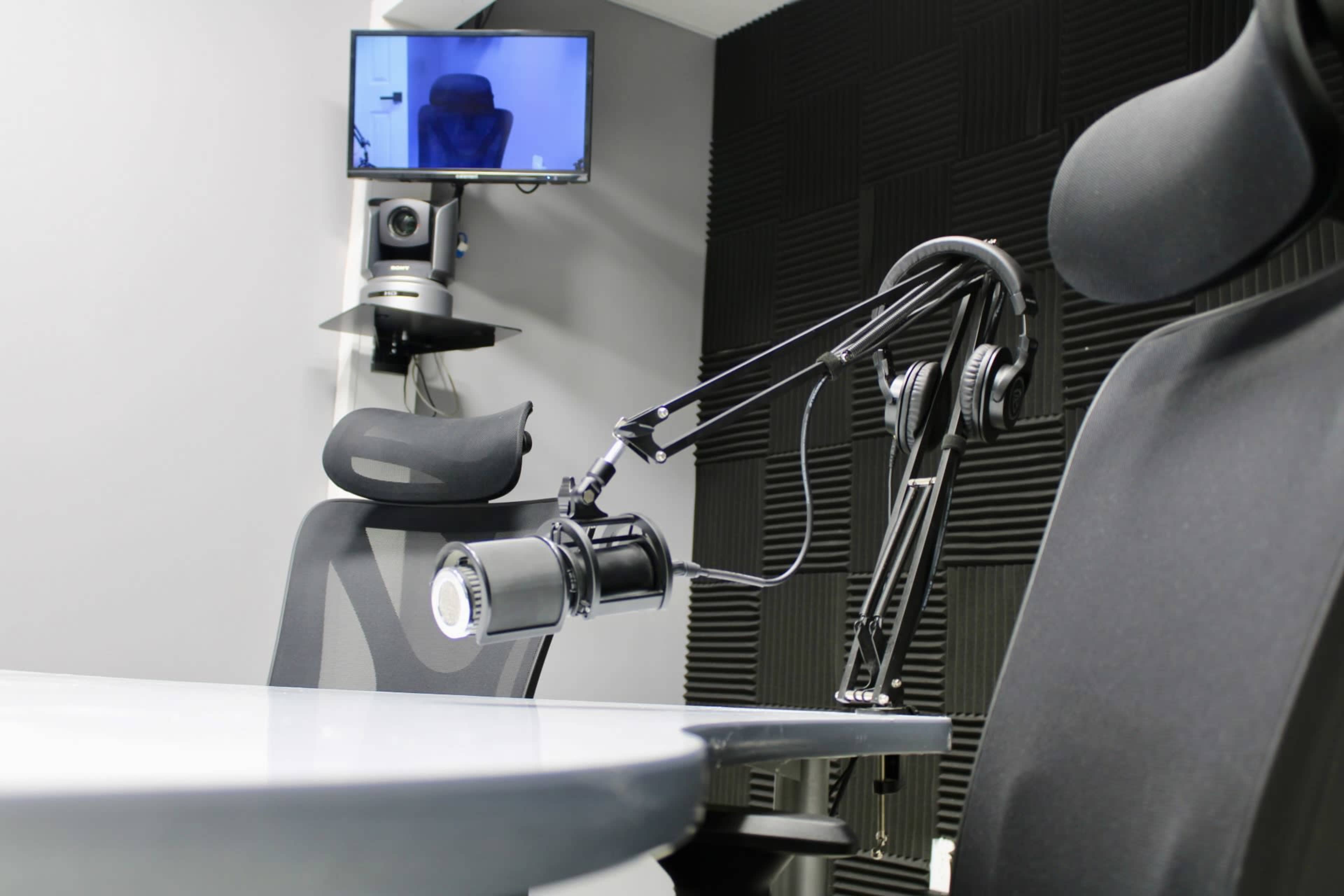 The image shows a recording studio setup with a microphone on an adjustable arm, a modern office chair, and a small monitor mounted on the wall.