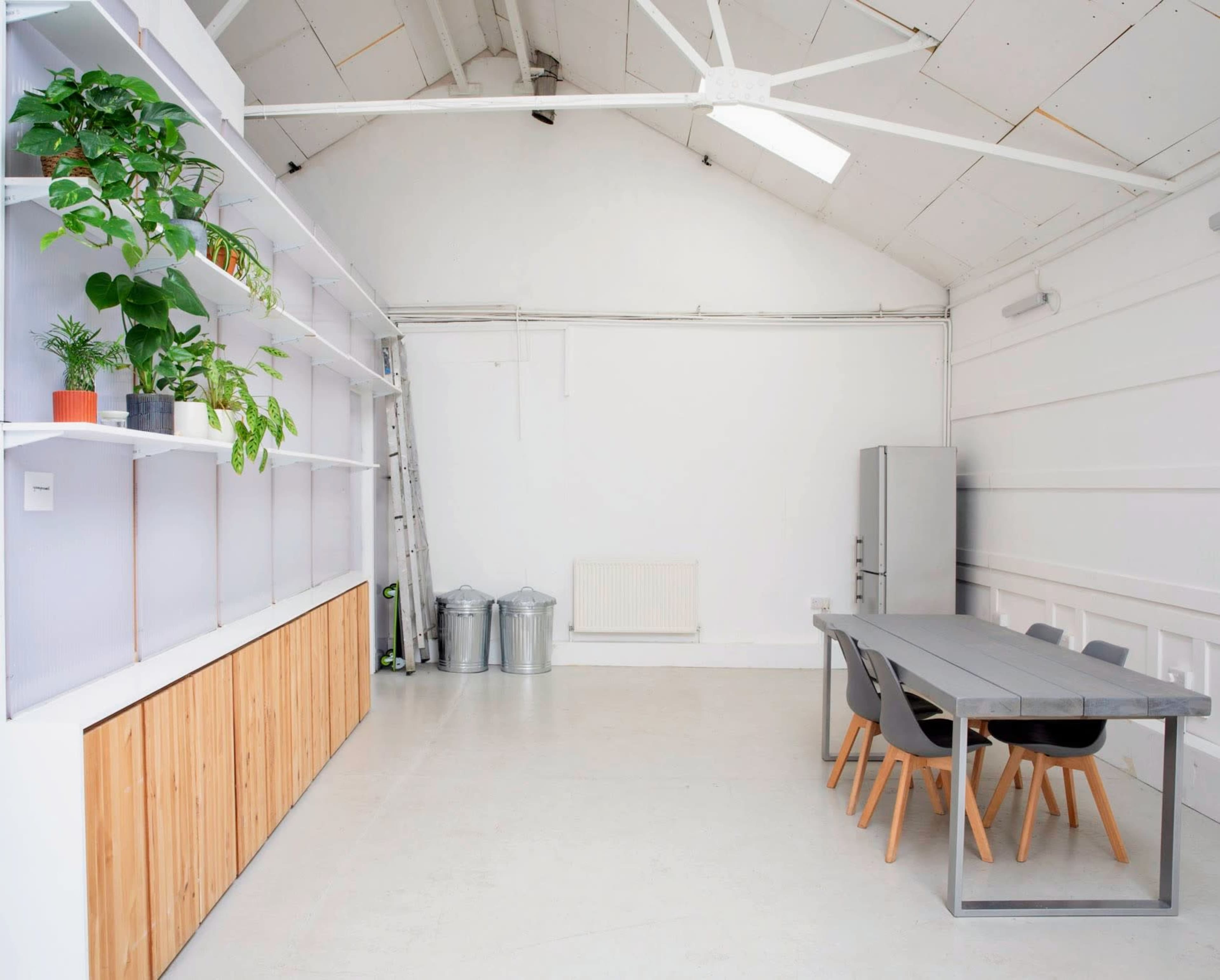 The image shows a spacious, minimalist interior with a long dining table, gray chairs, and a wooden storage unit against a white wall, complemented by potted plants on shelves.