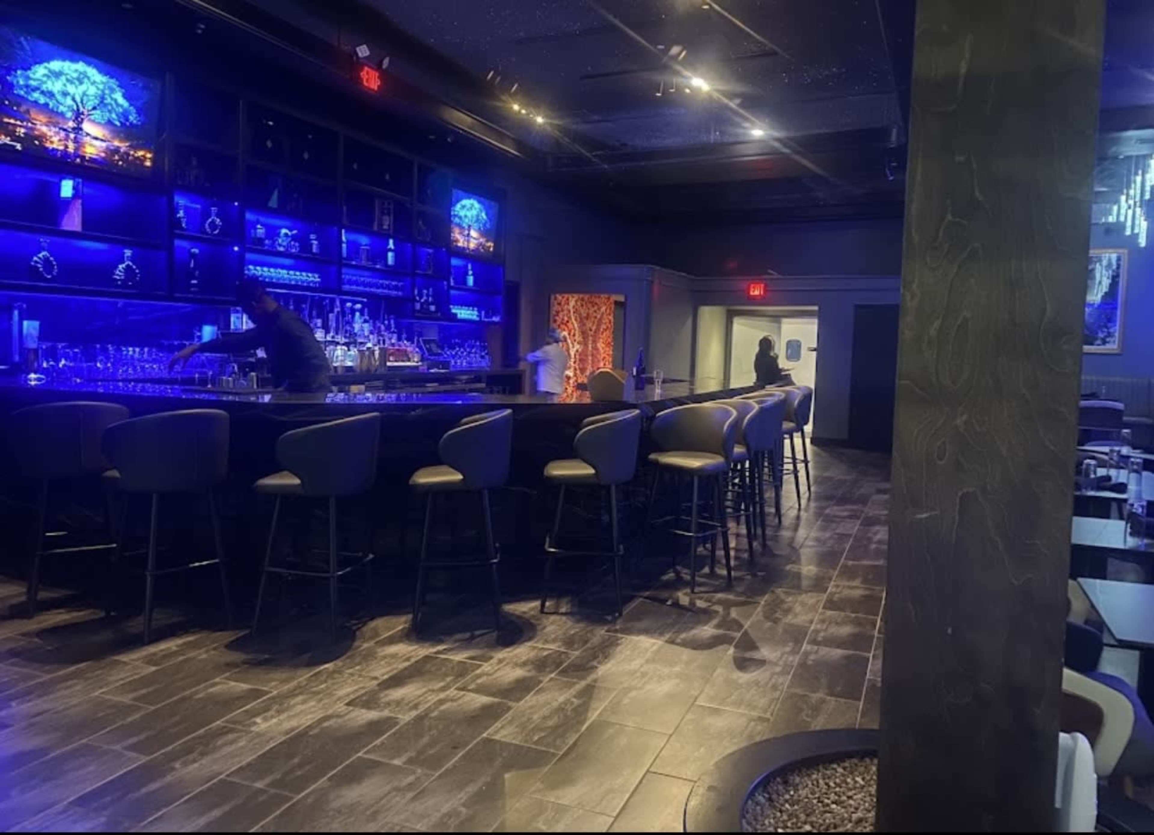 The image shows a dimly lit bar area with high stools along a curved counter and shelves displaying illuminated bottles.
