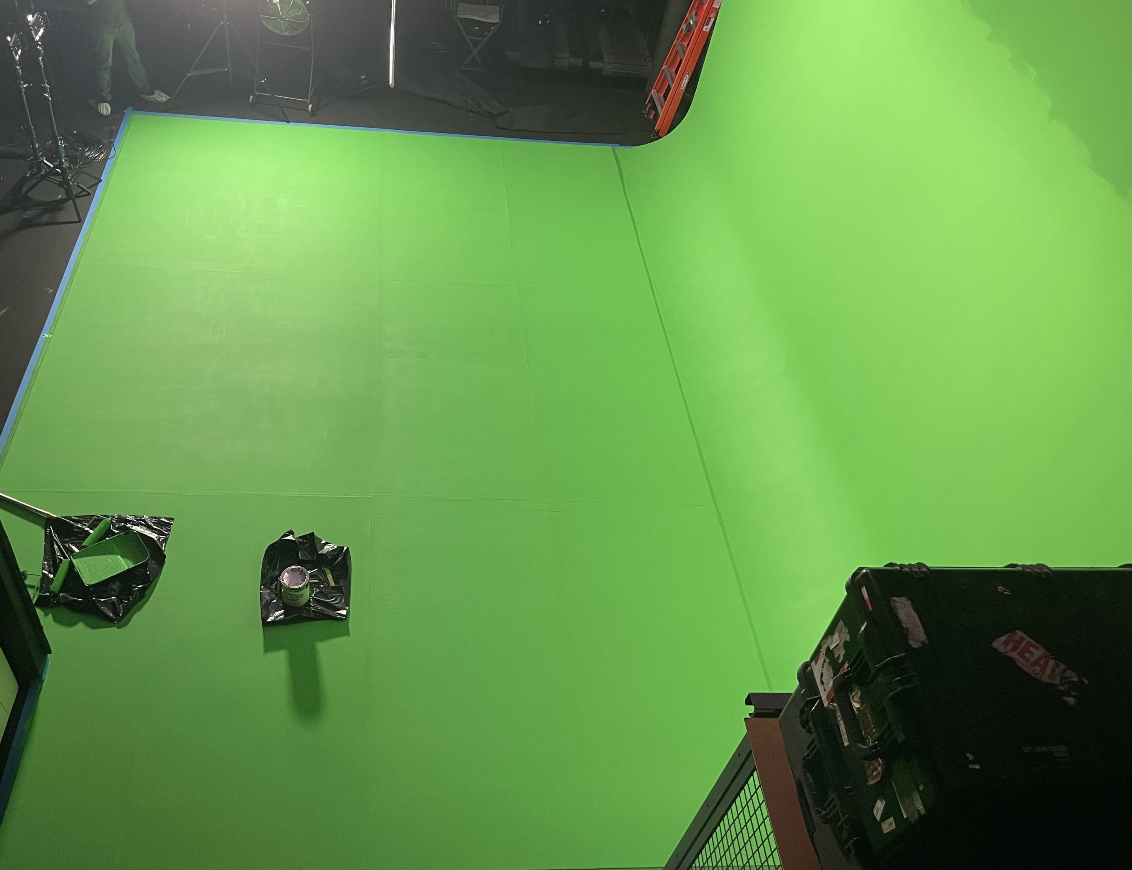 The image shows a green screen studio setup with a large green backdrop and some equipment on the floor.