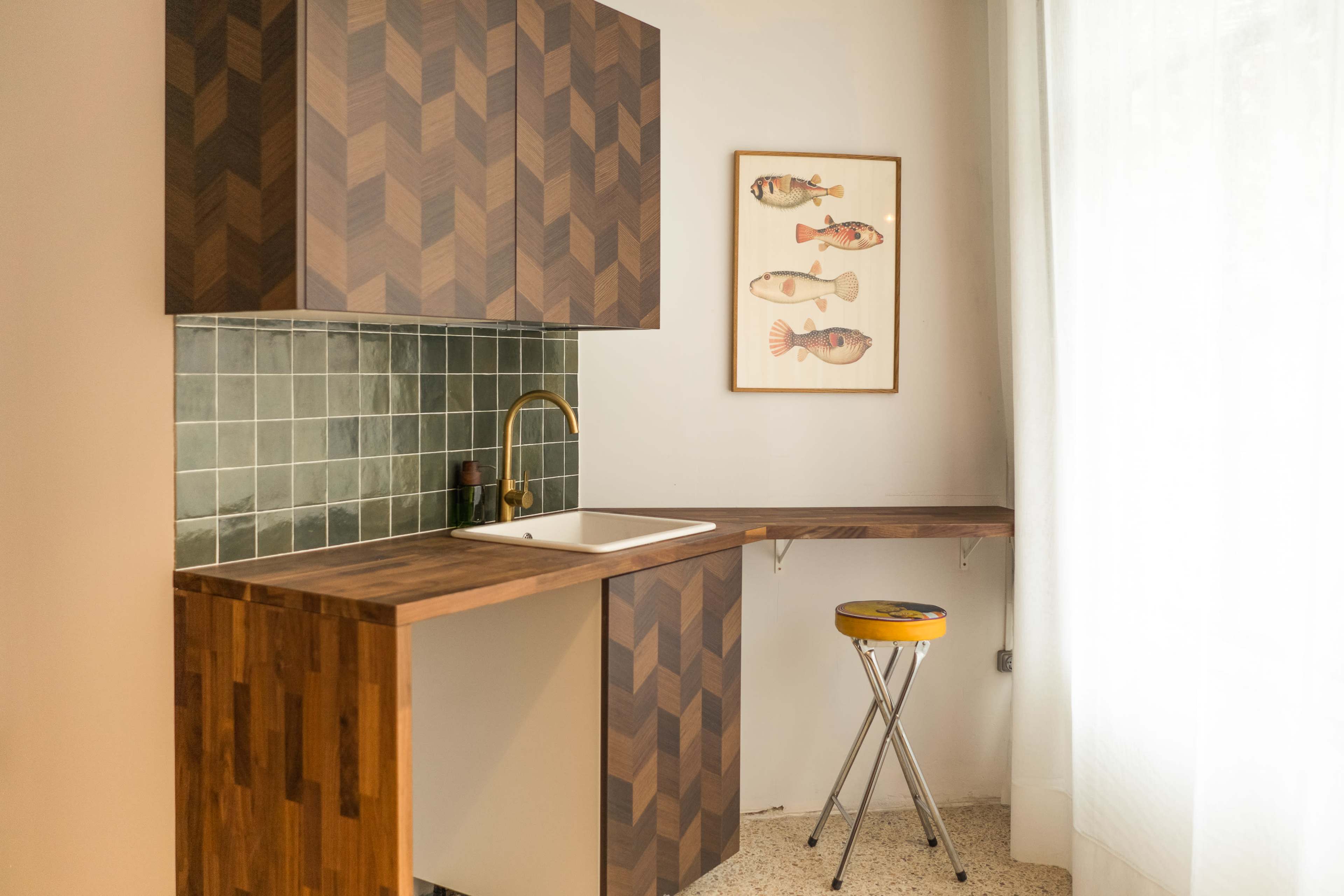 A corner kitchen with a wooden countertop, a white sink, green tiled backsplash, and a framed fish illustration on the wall.