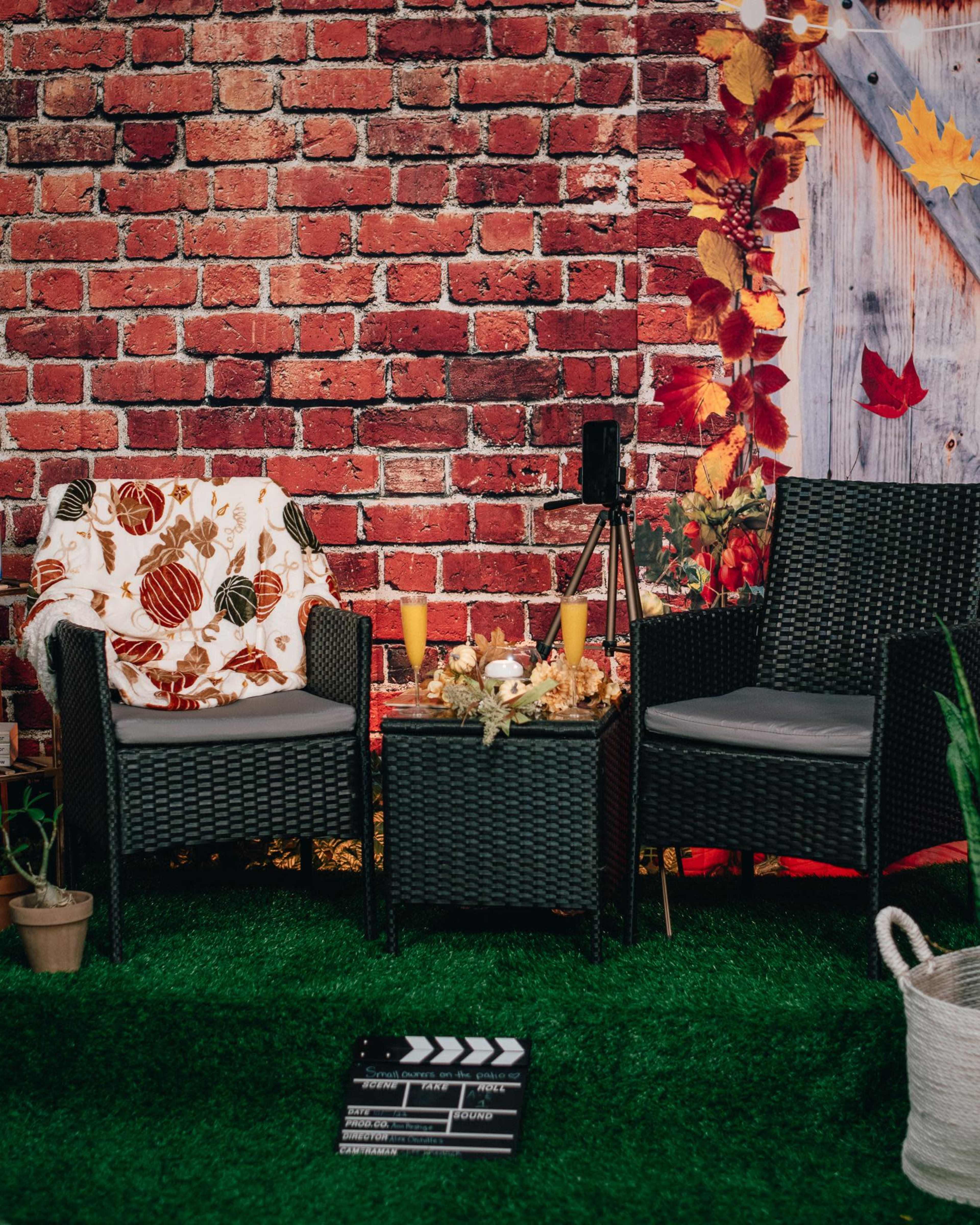 The scene features two black wicker chairs with a small table between them, set against a brick wall adorned with autumn decor, a film clapperboard on the grass below.