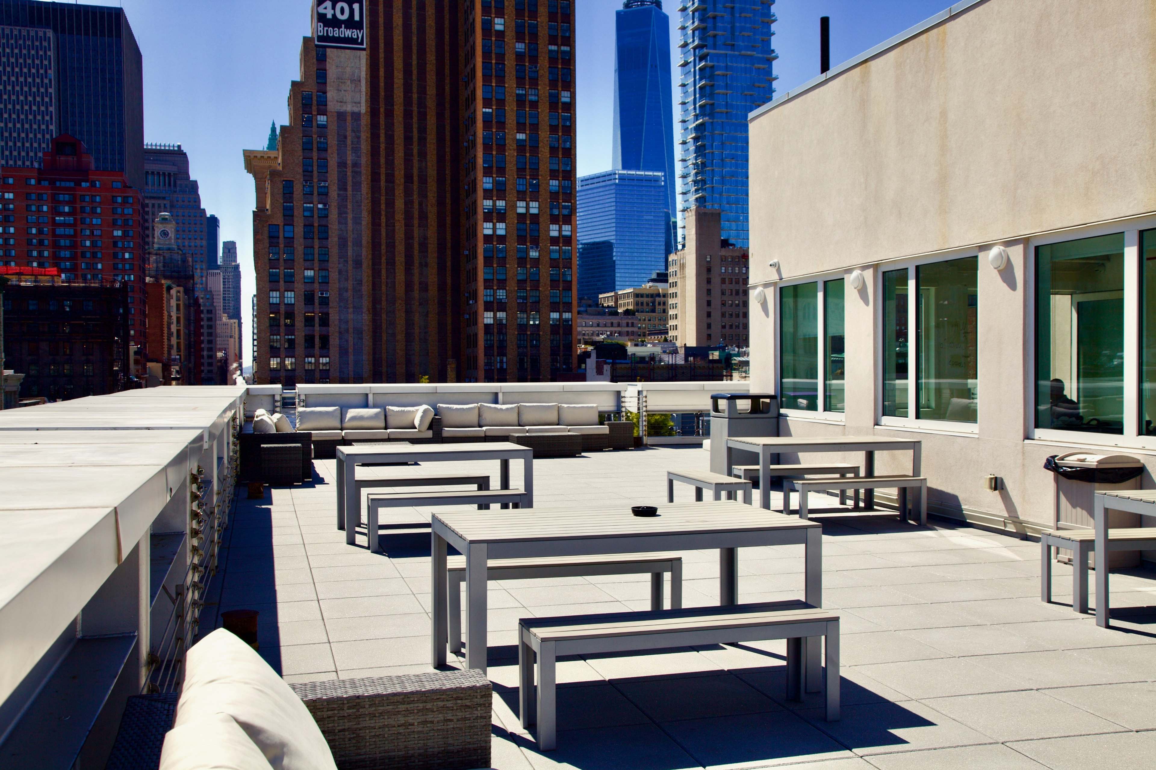 The image shows a rooftop terrace with several gray tables and benches, surrounded by tall buildings and a clear blue sky.