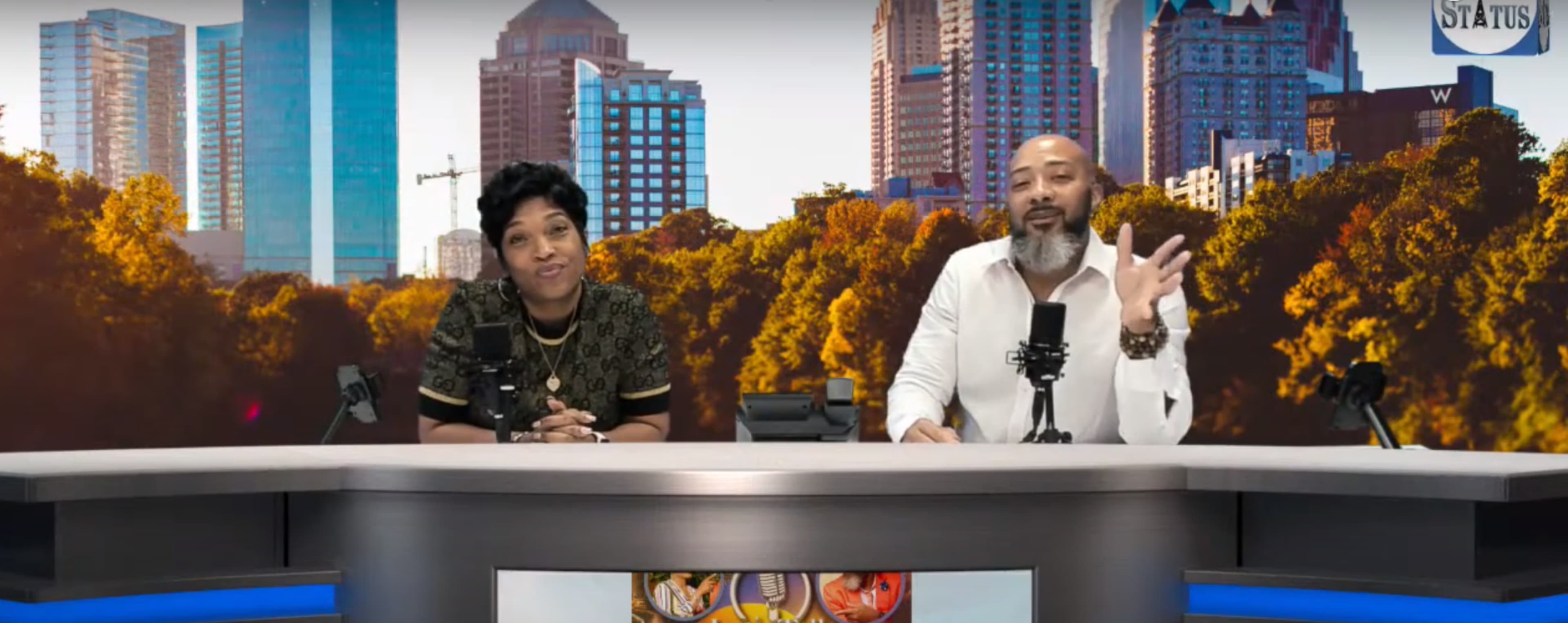 A man and a woman are seated at a desk with microphones in front of a backdrop featuring a city skyline and trees.