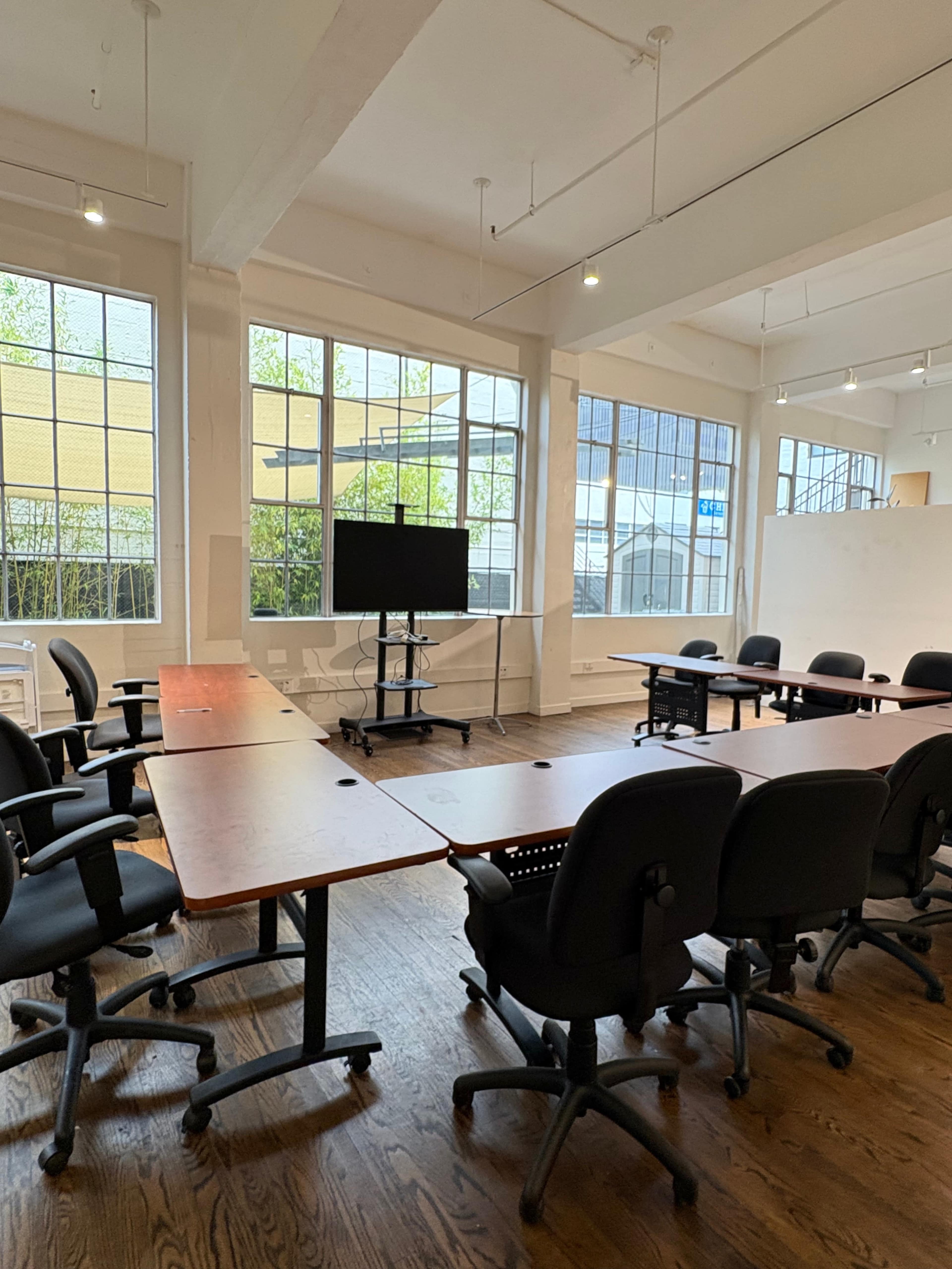 The image shows a spacious, well-lit conference room with multiple wooden tables arranged in a U-shape and several black office chairs.