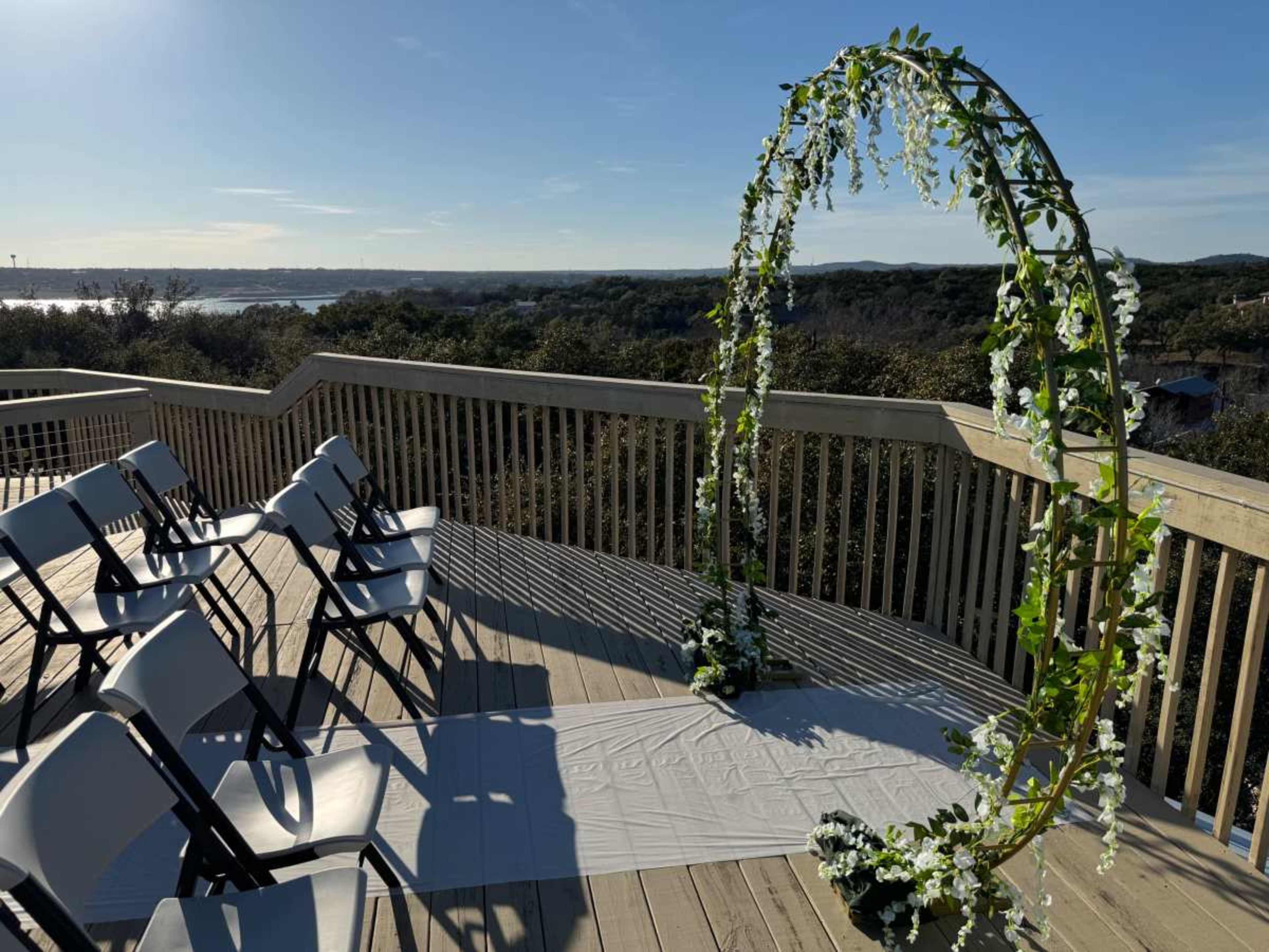 A row of white chairs is set up beside a floral-decorated arch on a wooden deck overlooking a scenic landscape.