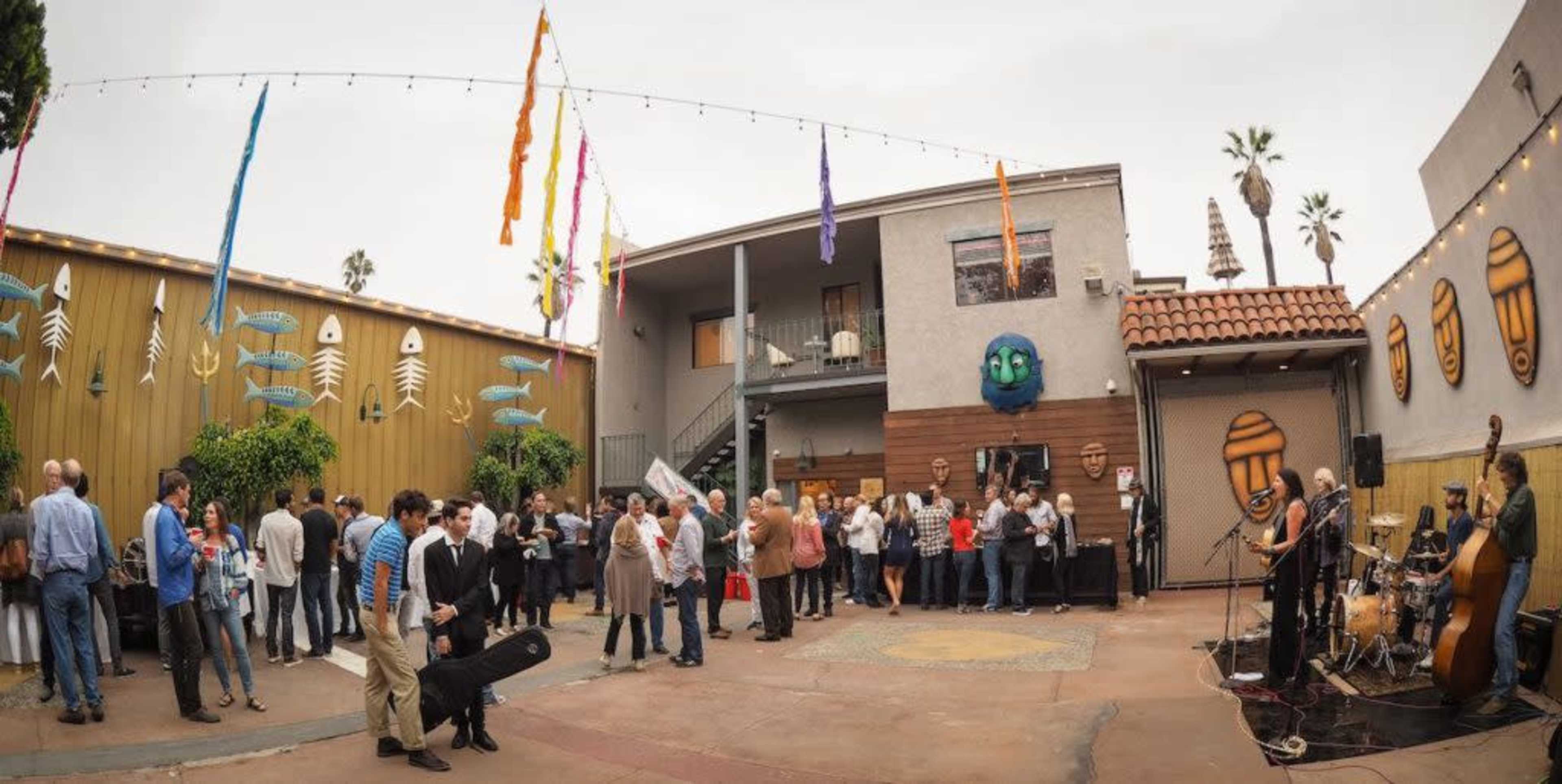 A crowd of people mingles in a courtyard featuring live music, colorful decorations, and palm trees.