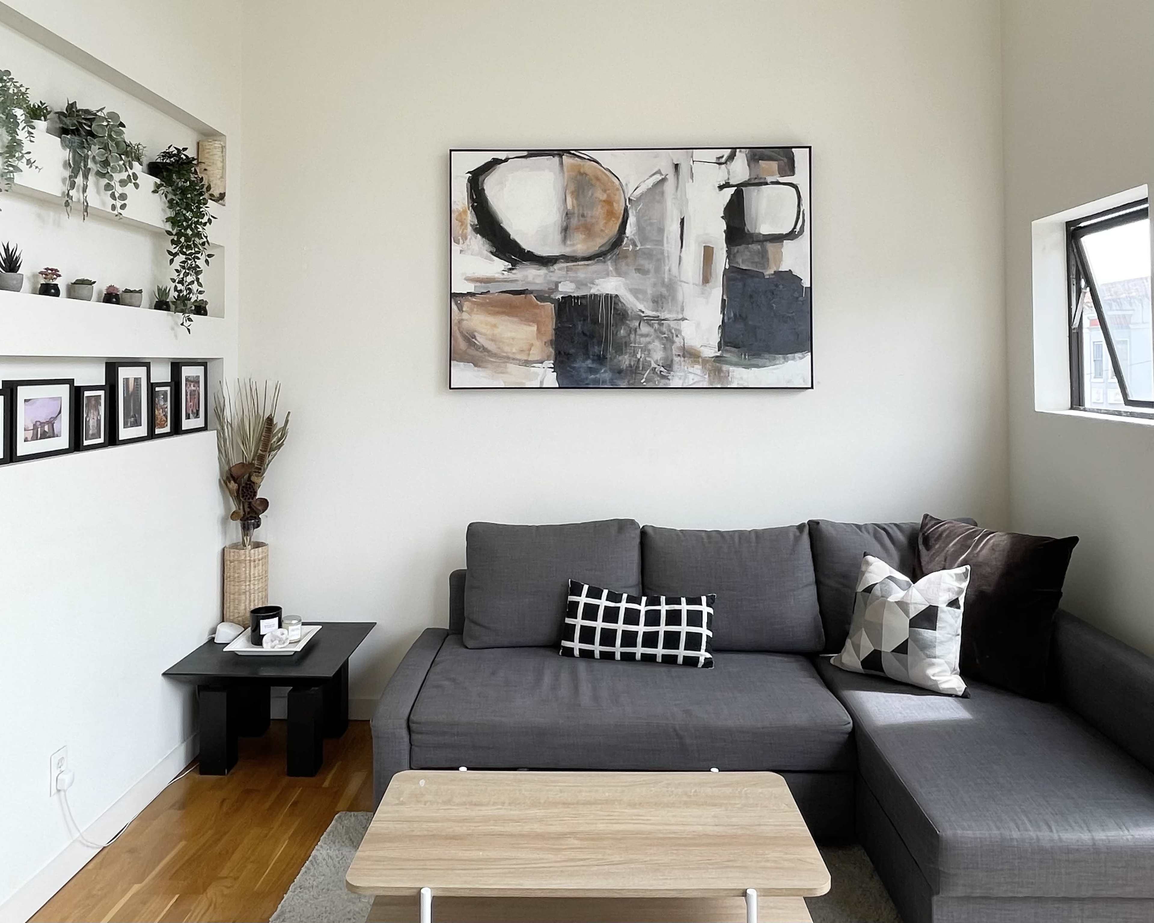 The image shows a modern living room featuring a gray sectional sofa, a wooden coffee table, and a wall-mounted artwork above a shelf with decorative plants.