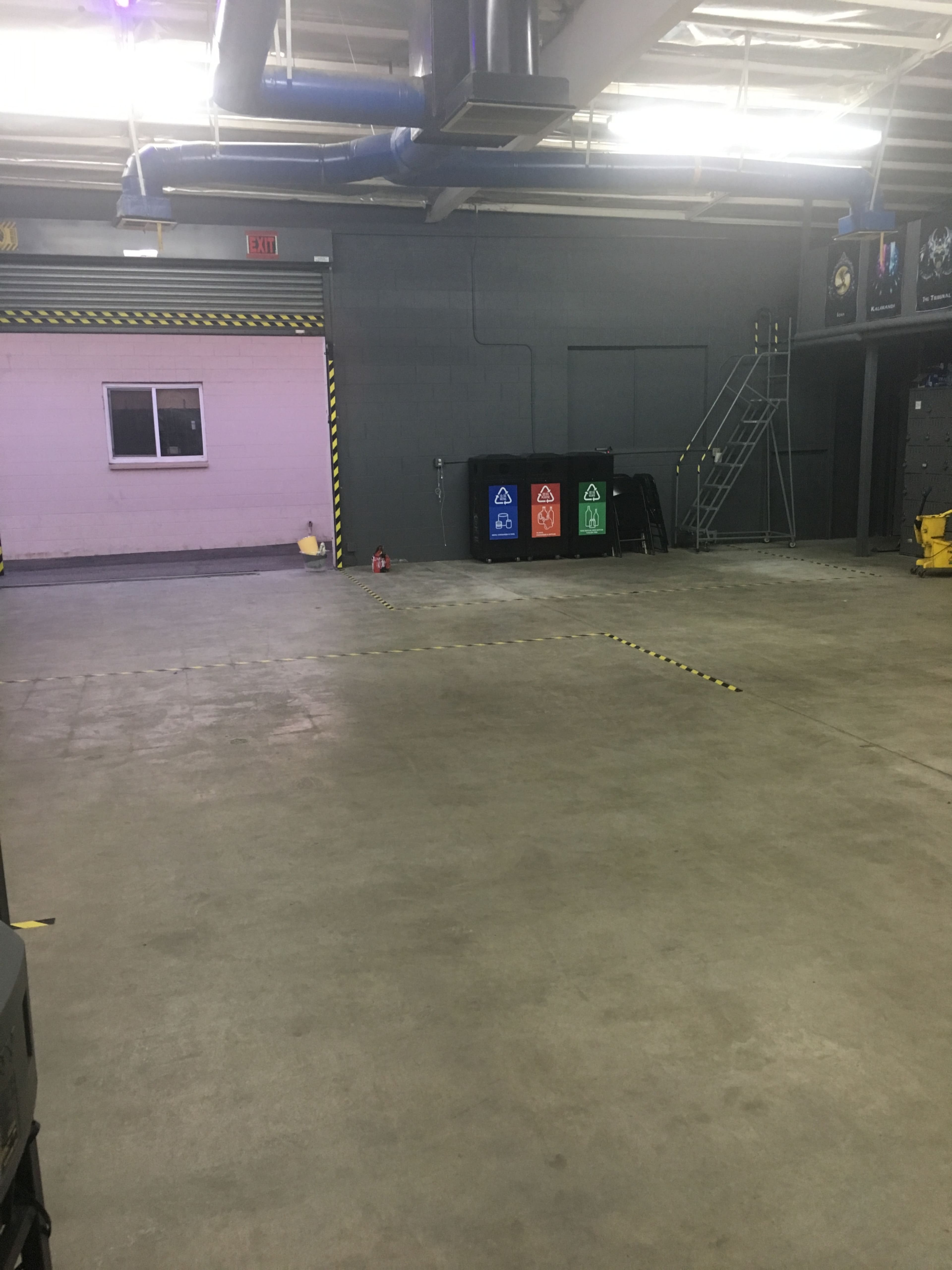 A large, empty warehouse or garage space with a concrete floor, a small window on the back wall, and recycling bins in three colors lined up against the wall.
