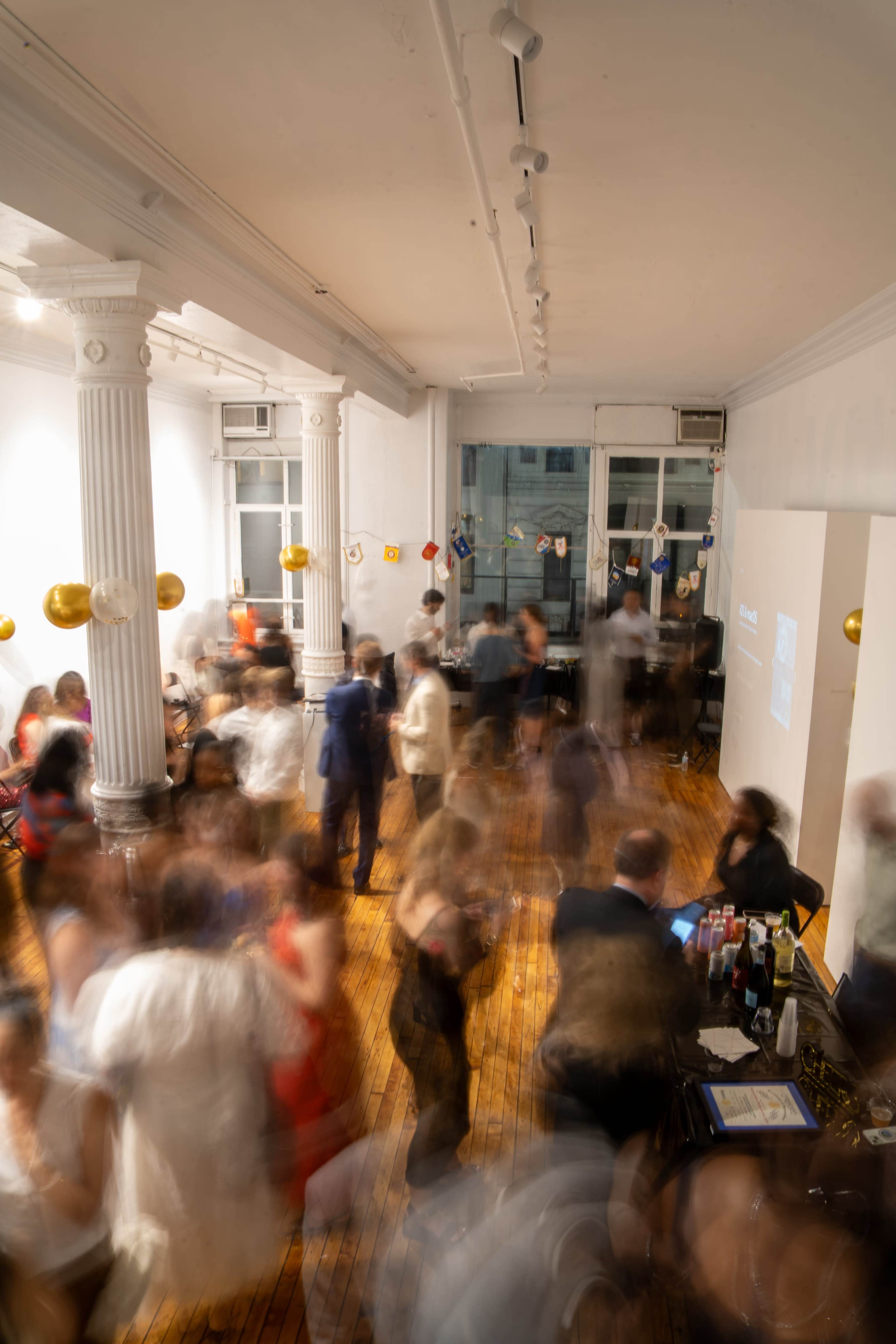 A crowd of people mingles in a spacious room adorned with golden balloons and large windows, creating a lively atmosphere.