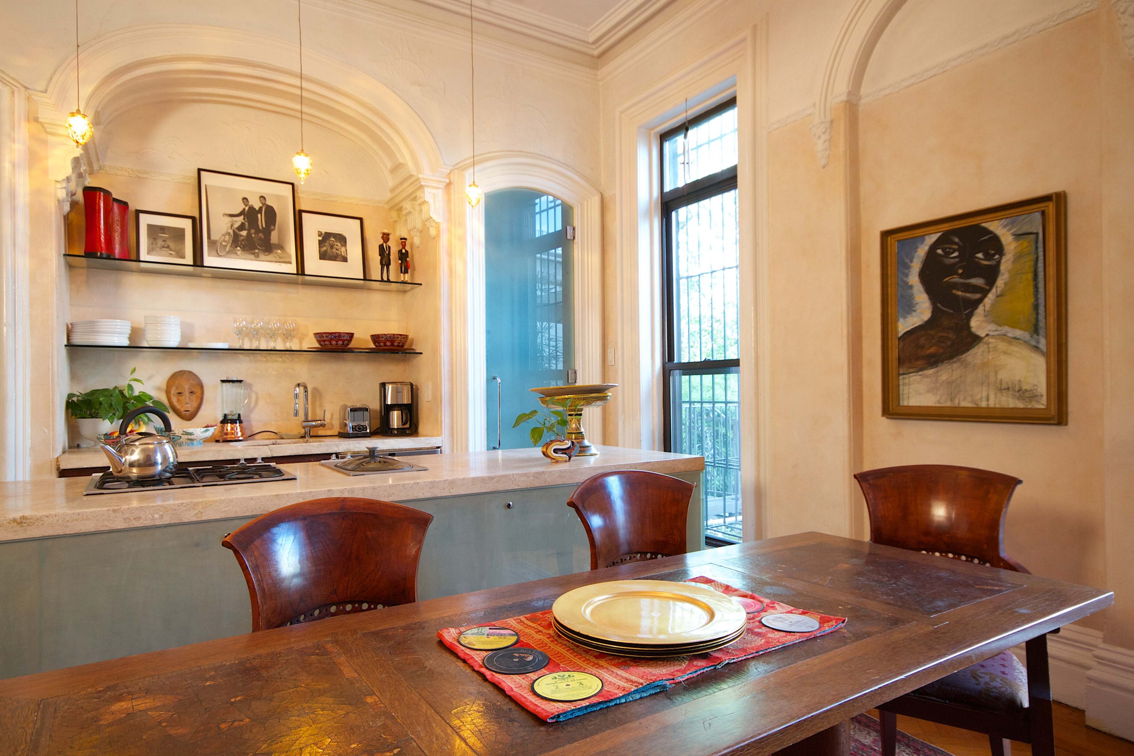 A kitchen and dining area feature a wooden table set with plates, a shelf of kitchen items, and an art piece on the wall.