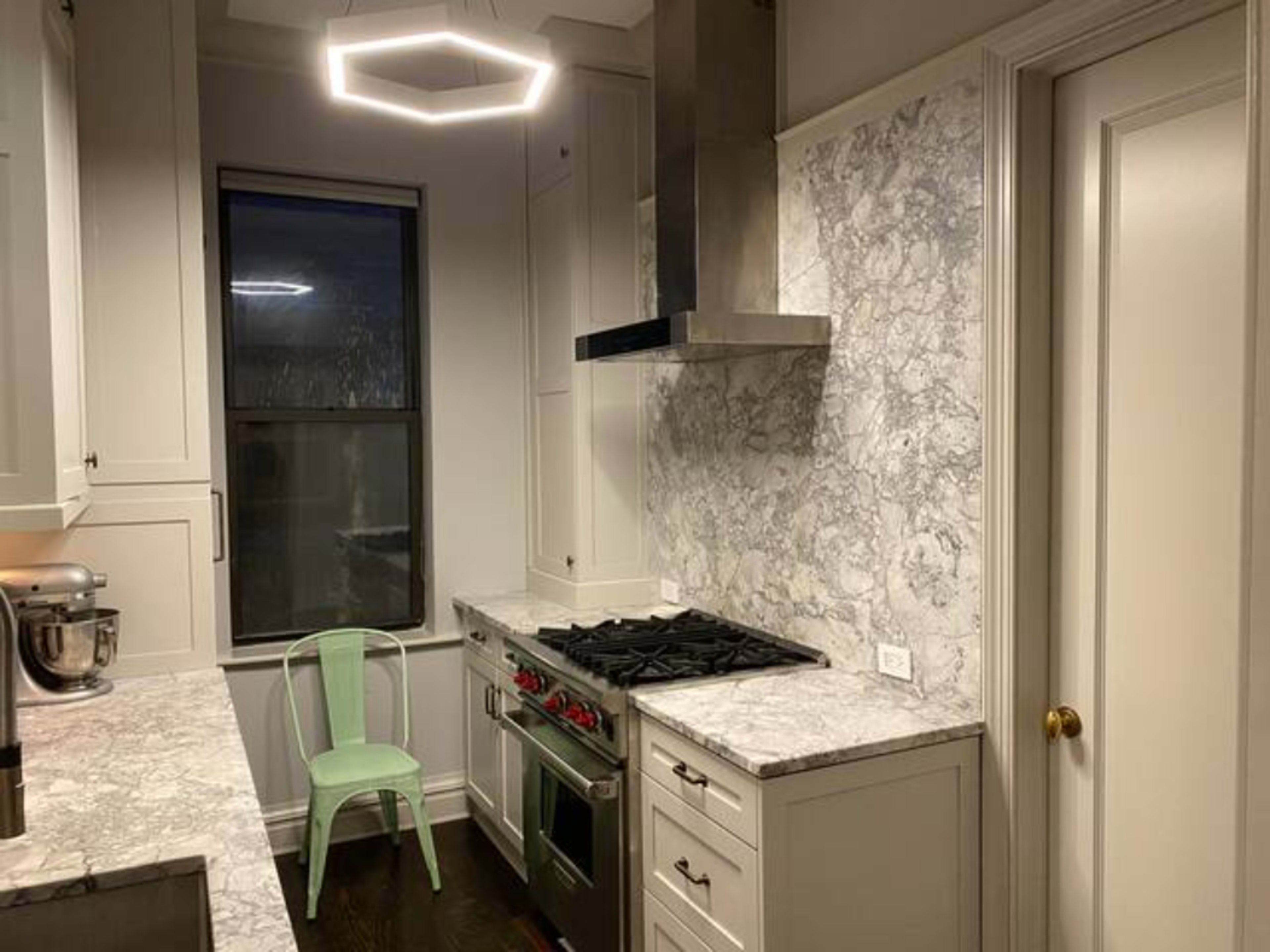 A modern kitchen features gray marble countertops, a stainless steel range hood, a green chair, and a hexagonal light fixture.