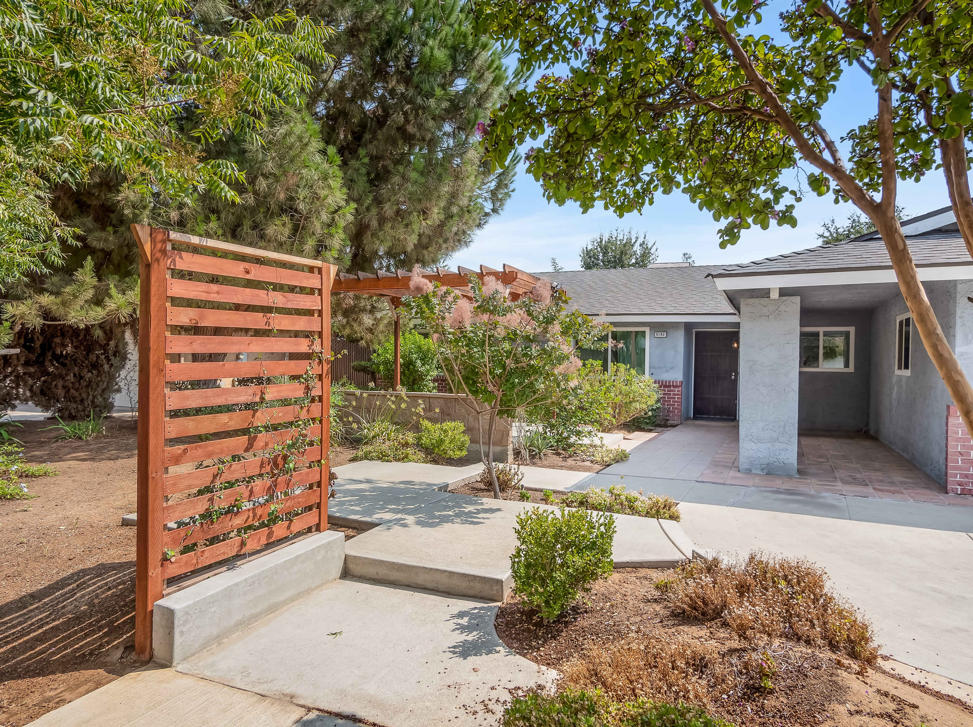 A landscaped entryway to a house featuring a wooden trellis, pathways, and greenery.