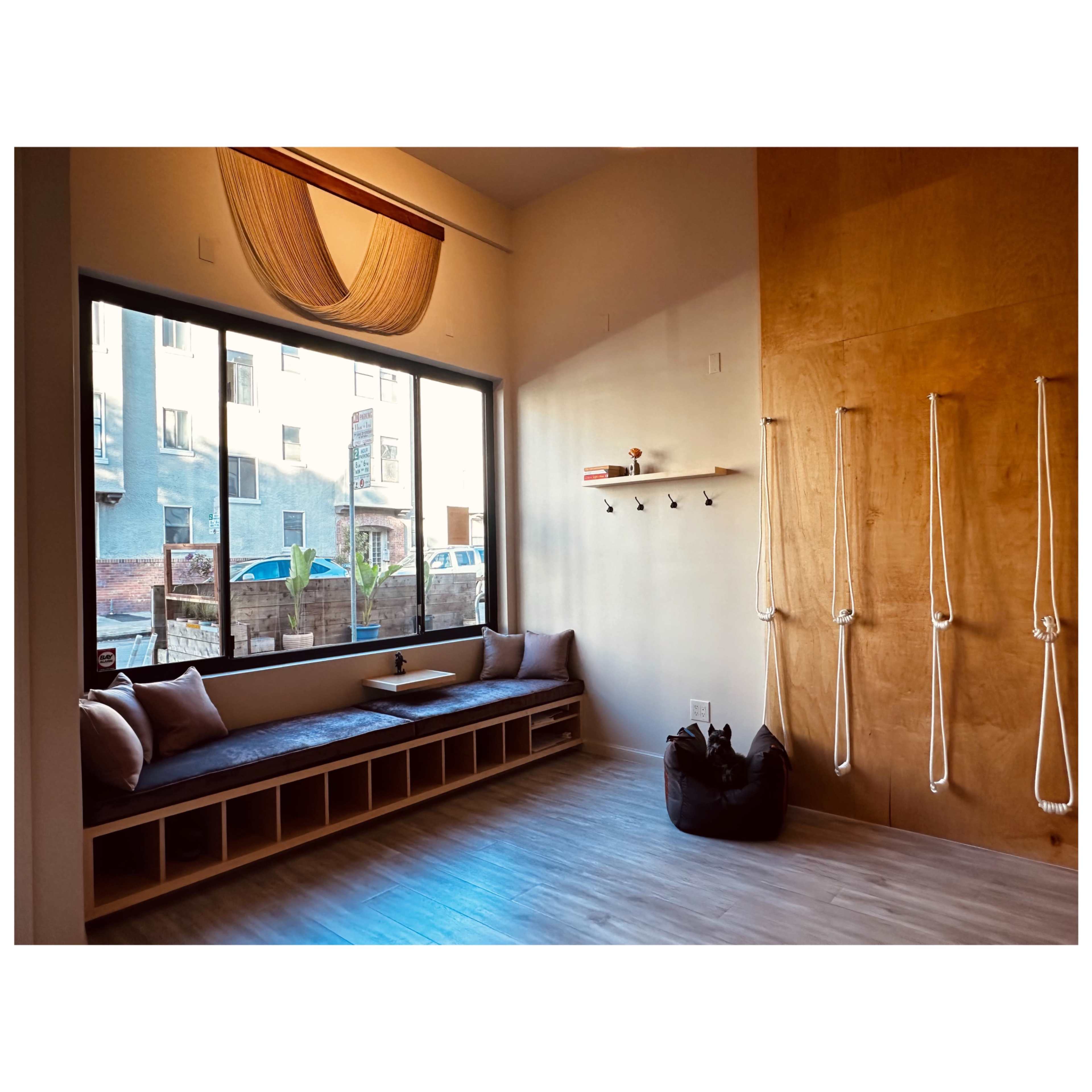 The image shows a minimalist yoga or meditation room with a large window, a built-in seating area, and wall ropes for stretching or support.