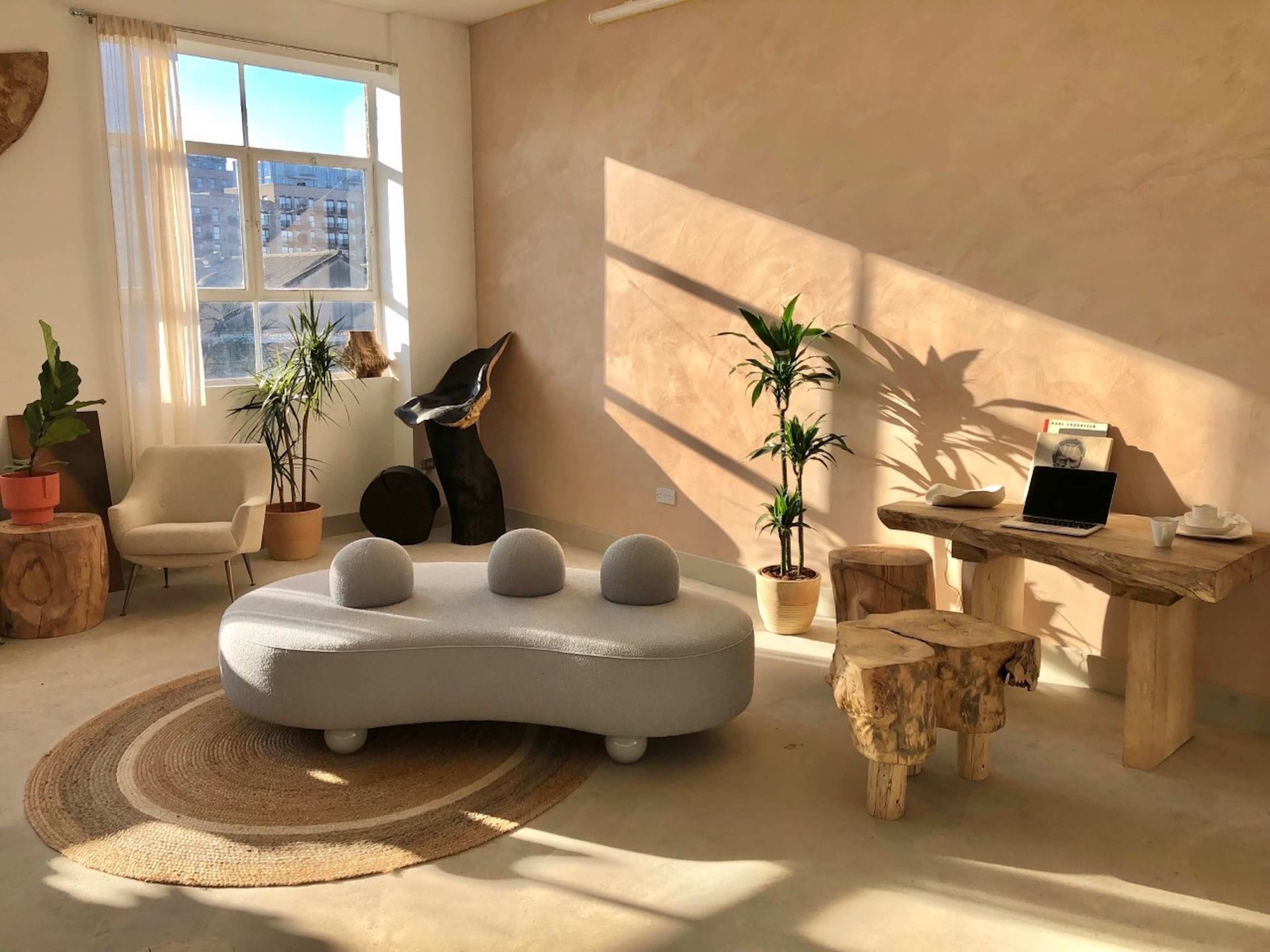 A modern, minimalistic living space with a light-colored wall, a circular gray sofa, natural wood furniture, and houseplants arranged beside a large window.