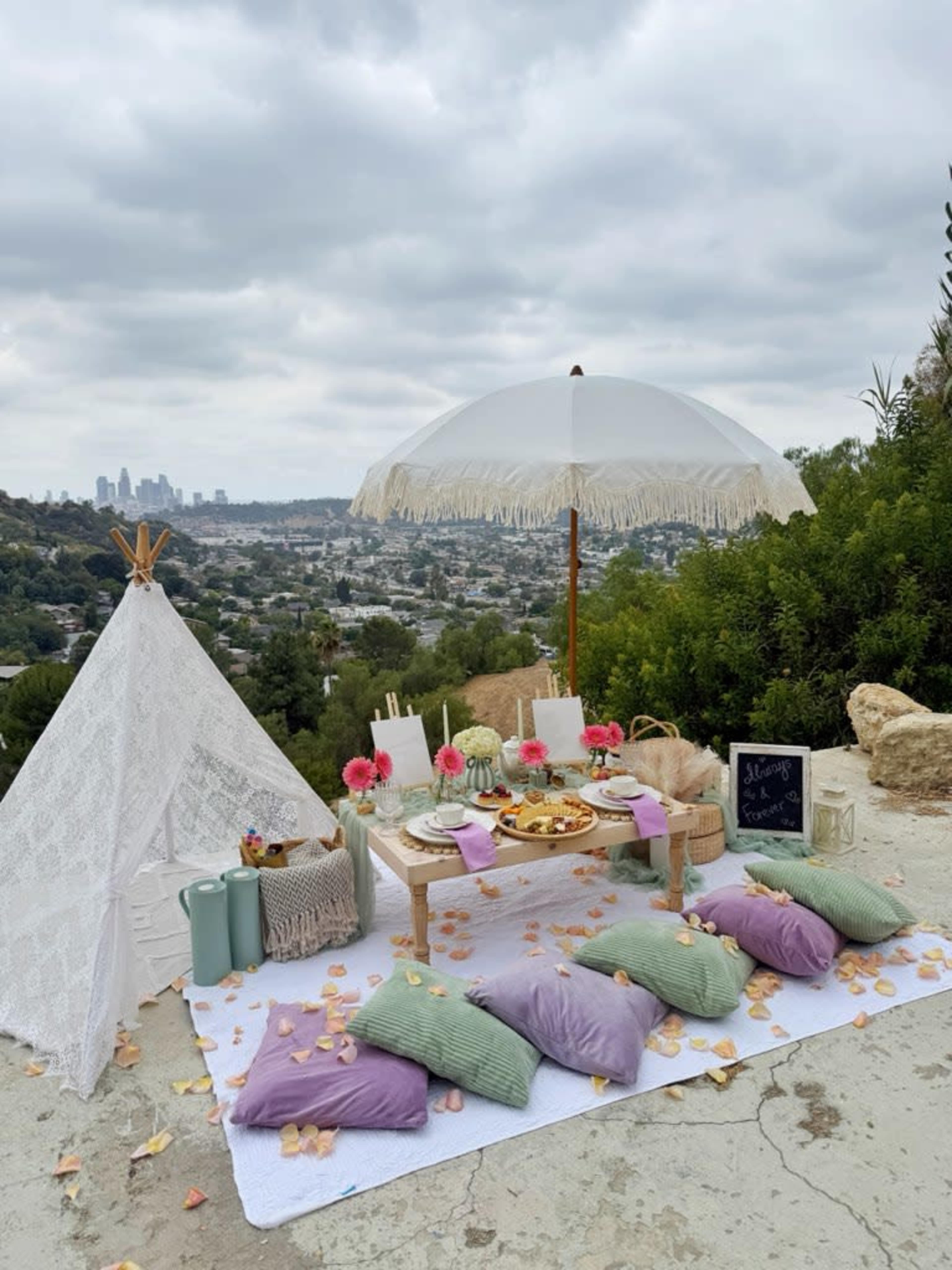 A picnic setup features a small table adorned with treats, colorful pillows, and a large parasol, set against a backdrop of city skyline and cloudy skies.