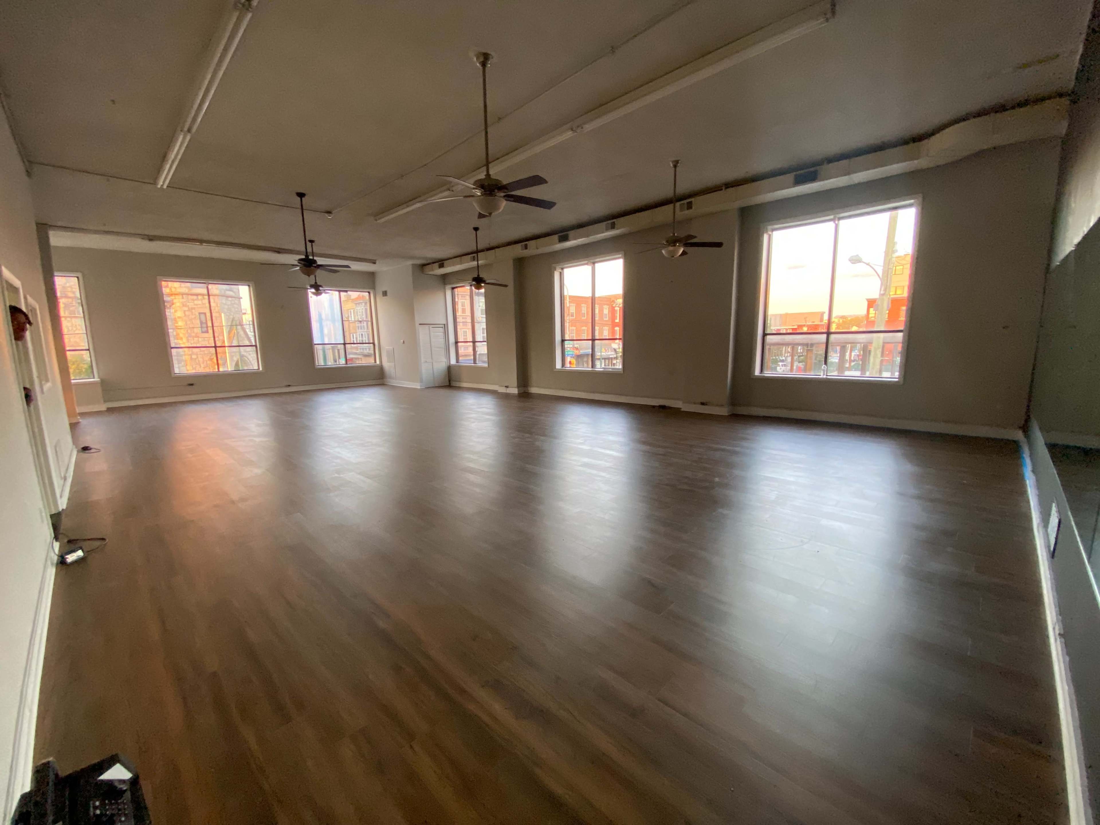 An empty room with wooden flooring, large windows, and ceiling fans.
