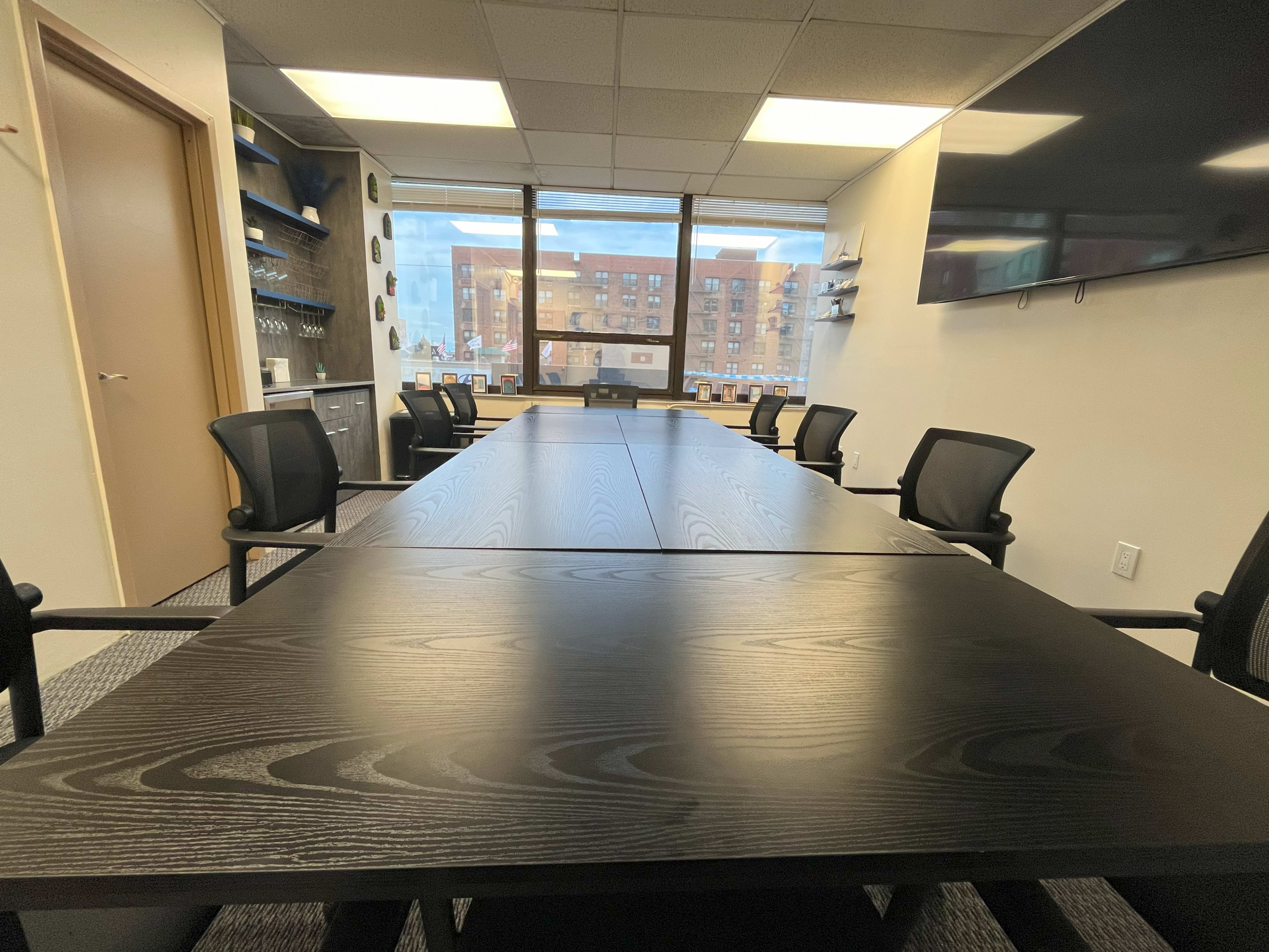 A conference room featuring a large wooden table surrounded by black chairs, with windows providing a view of a cityscape.