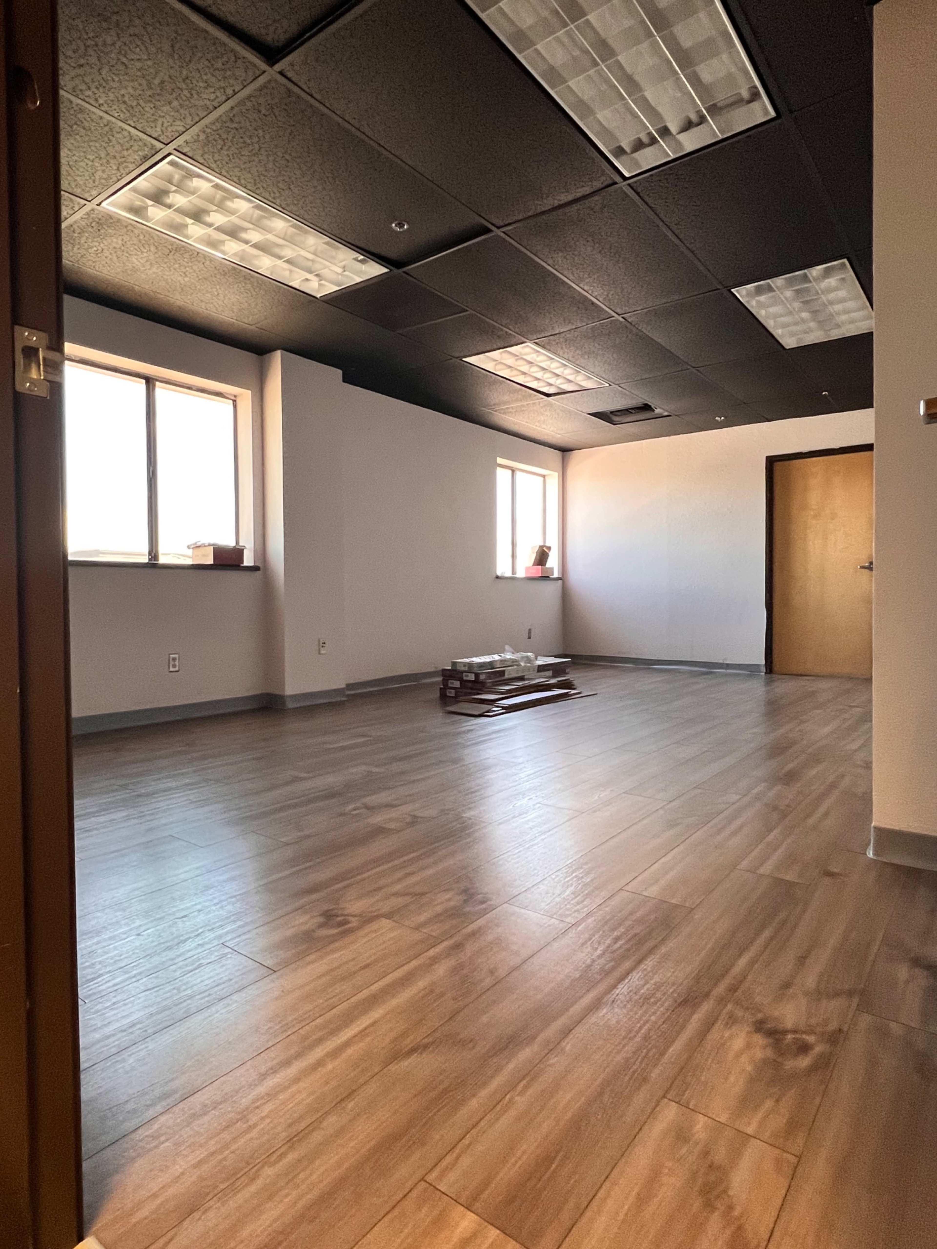 The image shows a vacant office space with wooden floors, two windows, and an open door leading into the room.