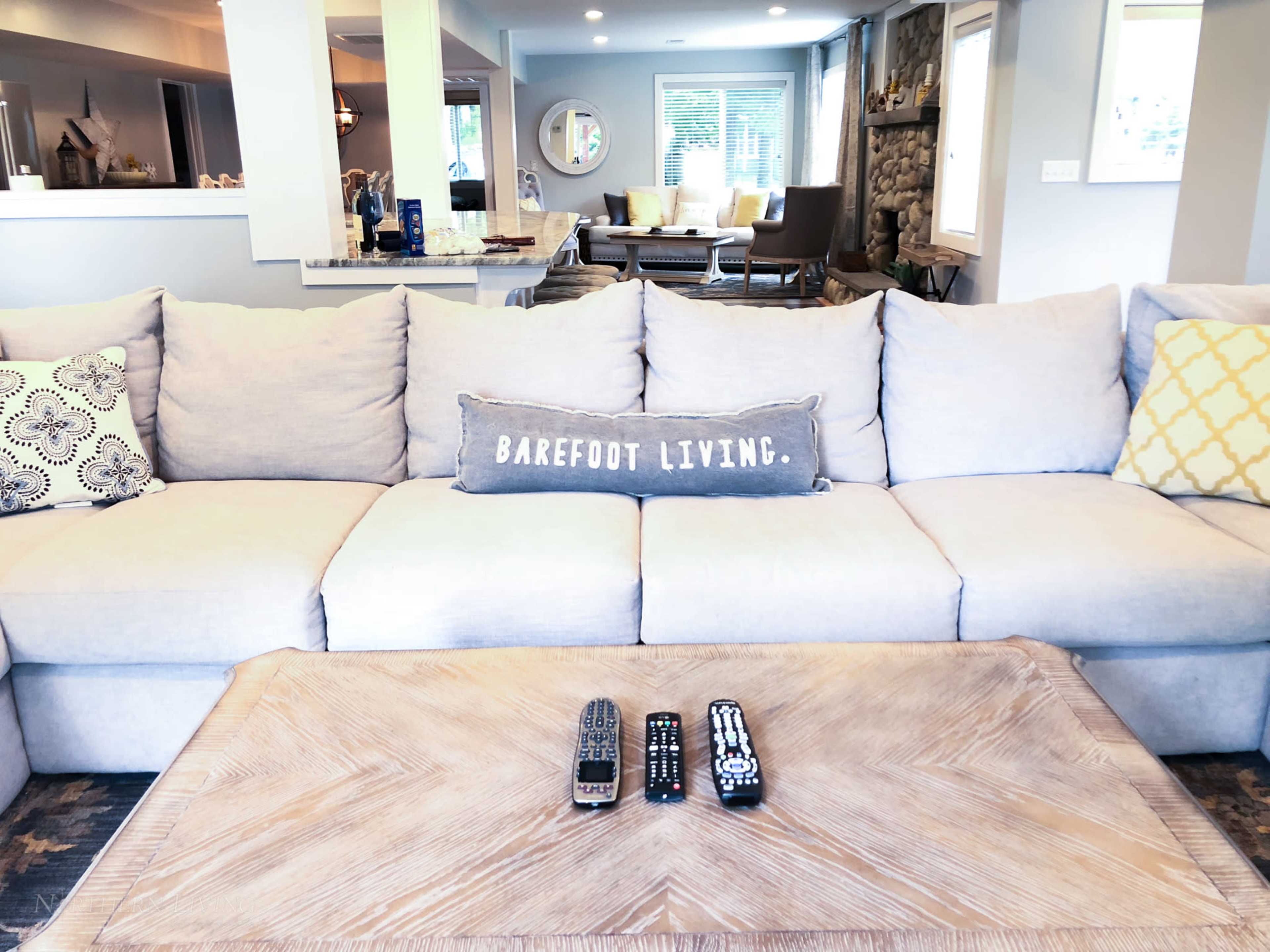 A light gray sectional sofa with decorative pillows is centered in a modern living room, accompanied by a wooden coffee table displaying remote controls.