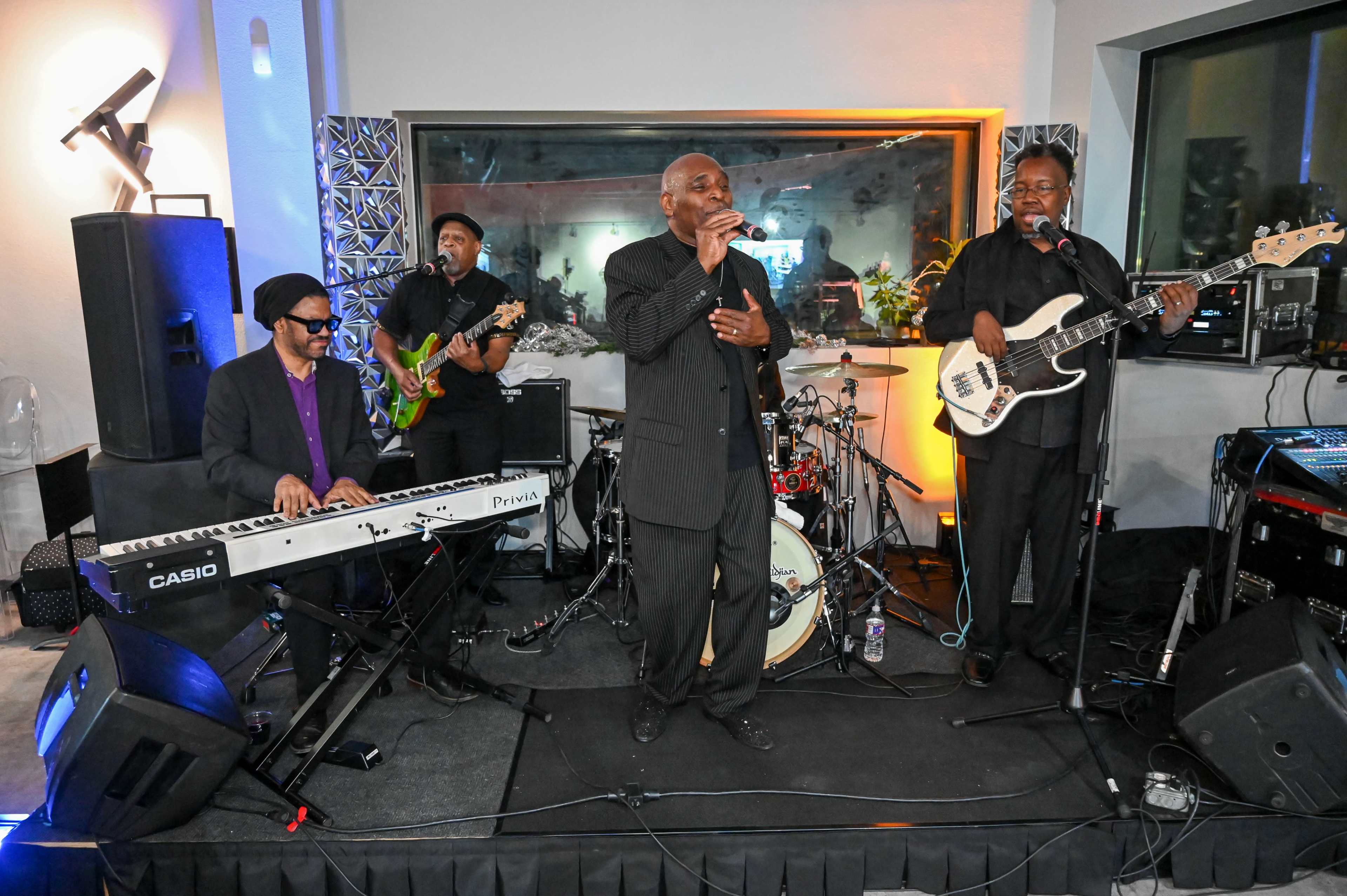 A musical group performs on stage with a keyboardist, guitarist, bassist, and a lead singer in a well-lit venue.