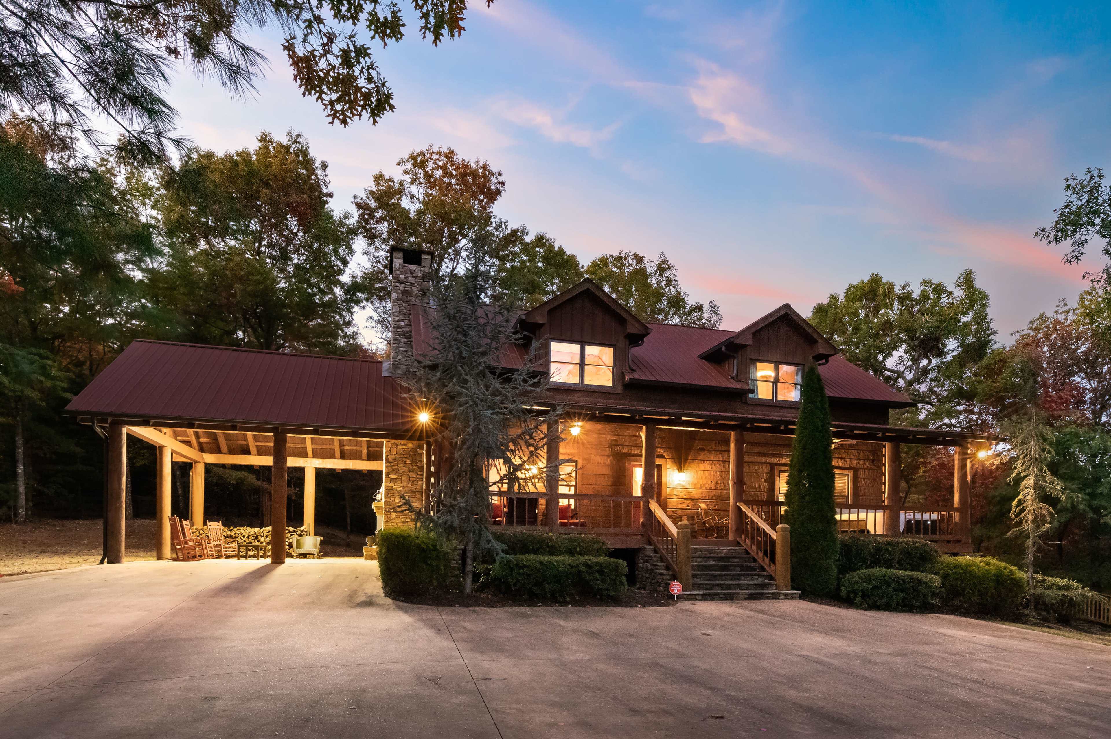 A rustic wooden house with a dark red metal roof and a wraparound porch stands in a wooded area at dusk.