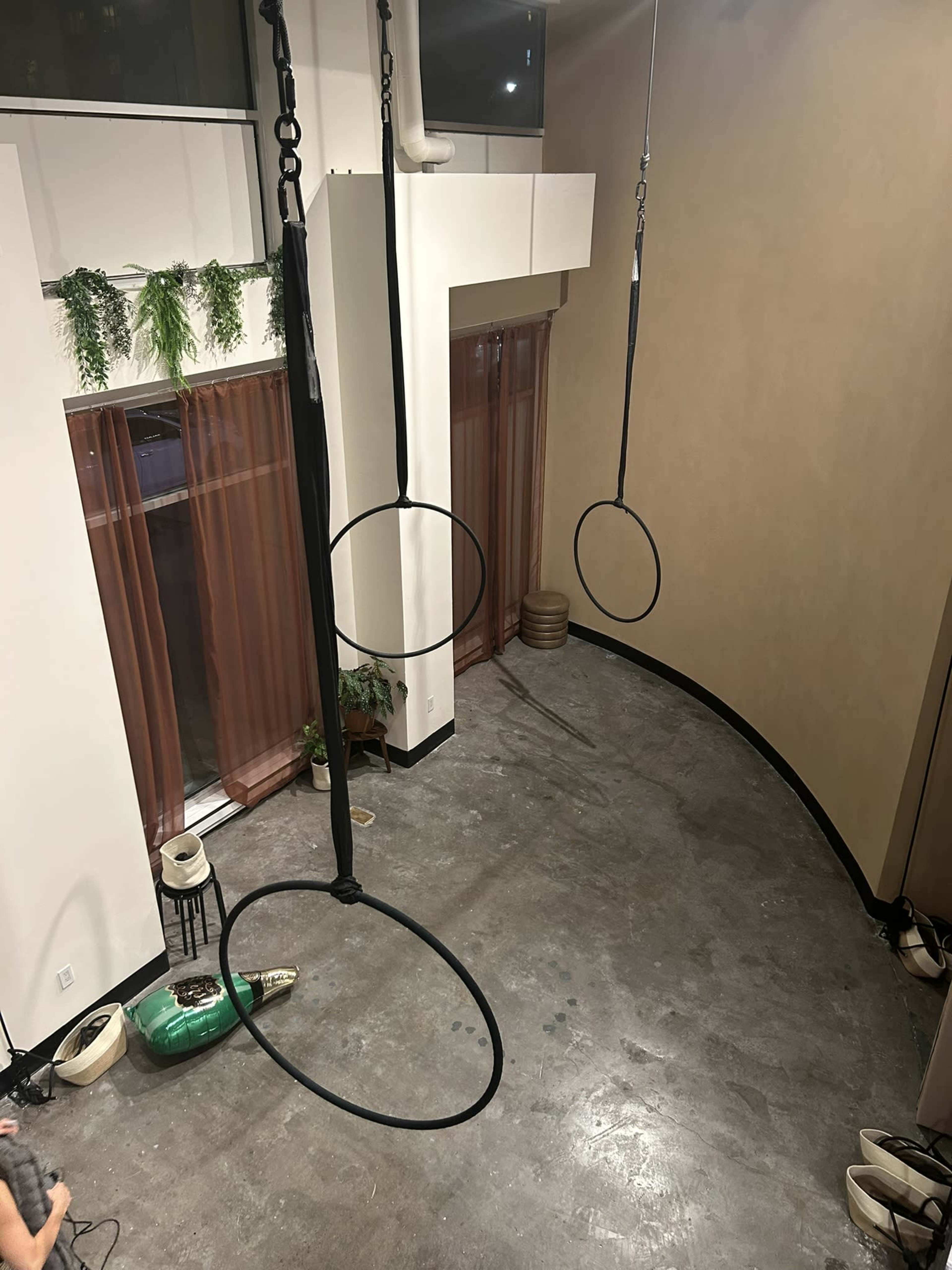 The image shows a room with three suspended hoops hanging from the ceiling, above a polished concrete floor, with large windows partially covered by curtains.