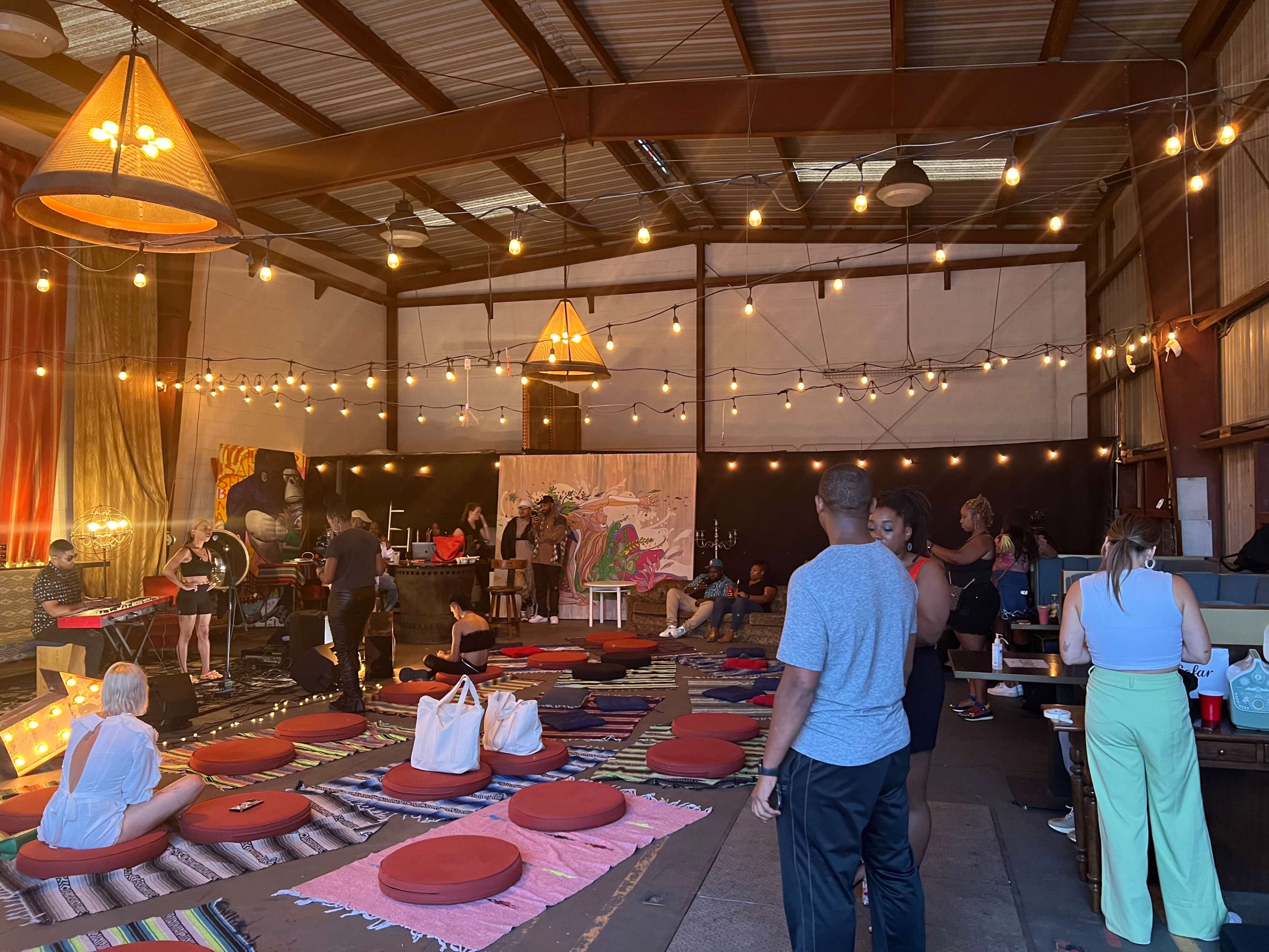 The image shows a lively indoor event space with people mingling, colorful floor cushions arranged on a patterned rug, and stage lights hanging above.
