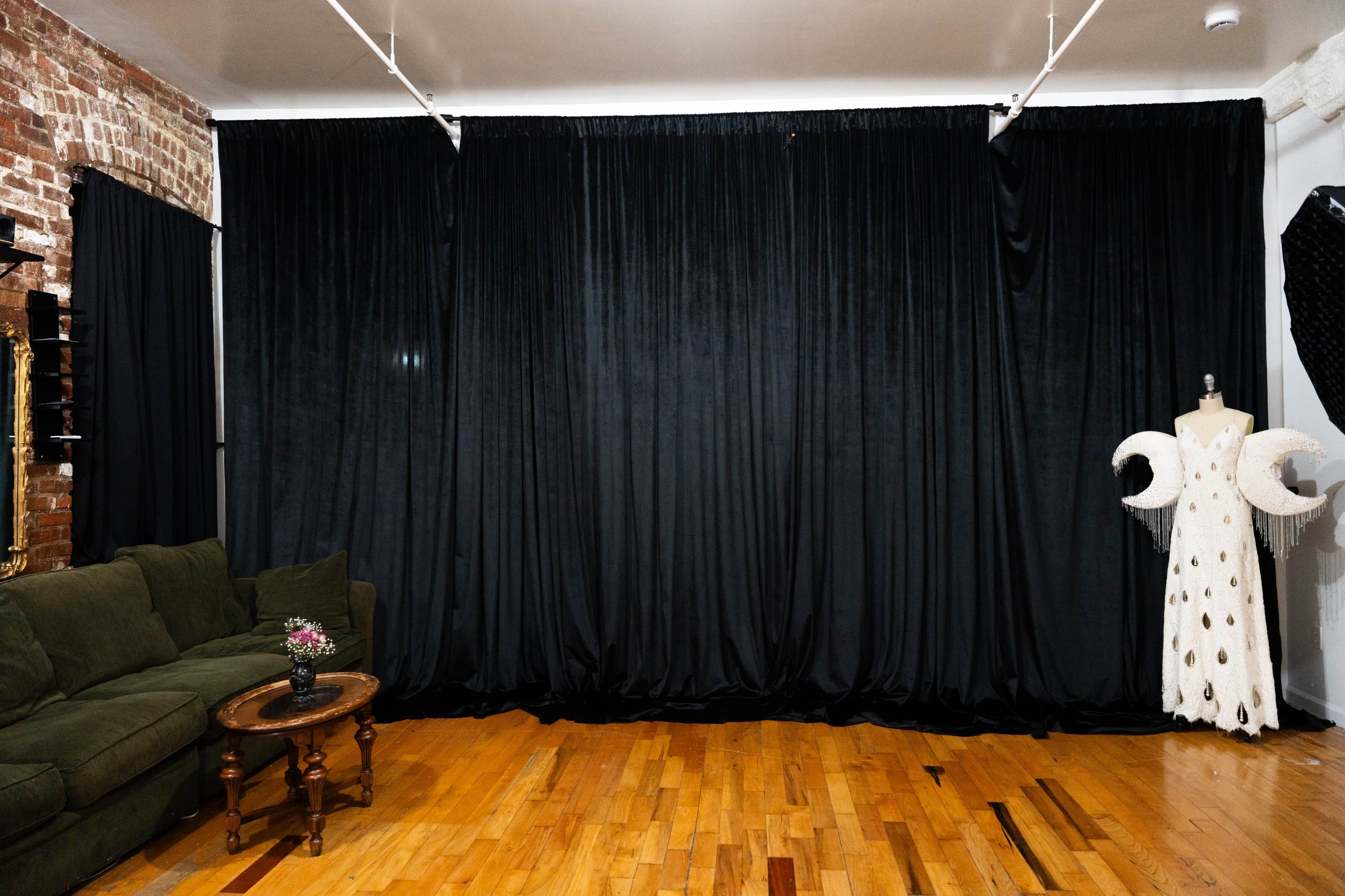 A room features a dark, textured curtain backdrop, a green sofa, a small flower vase on a wooden table, and a mannequin dressed in a white gown with decorative elements.