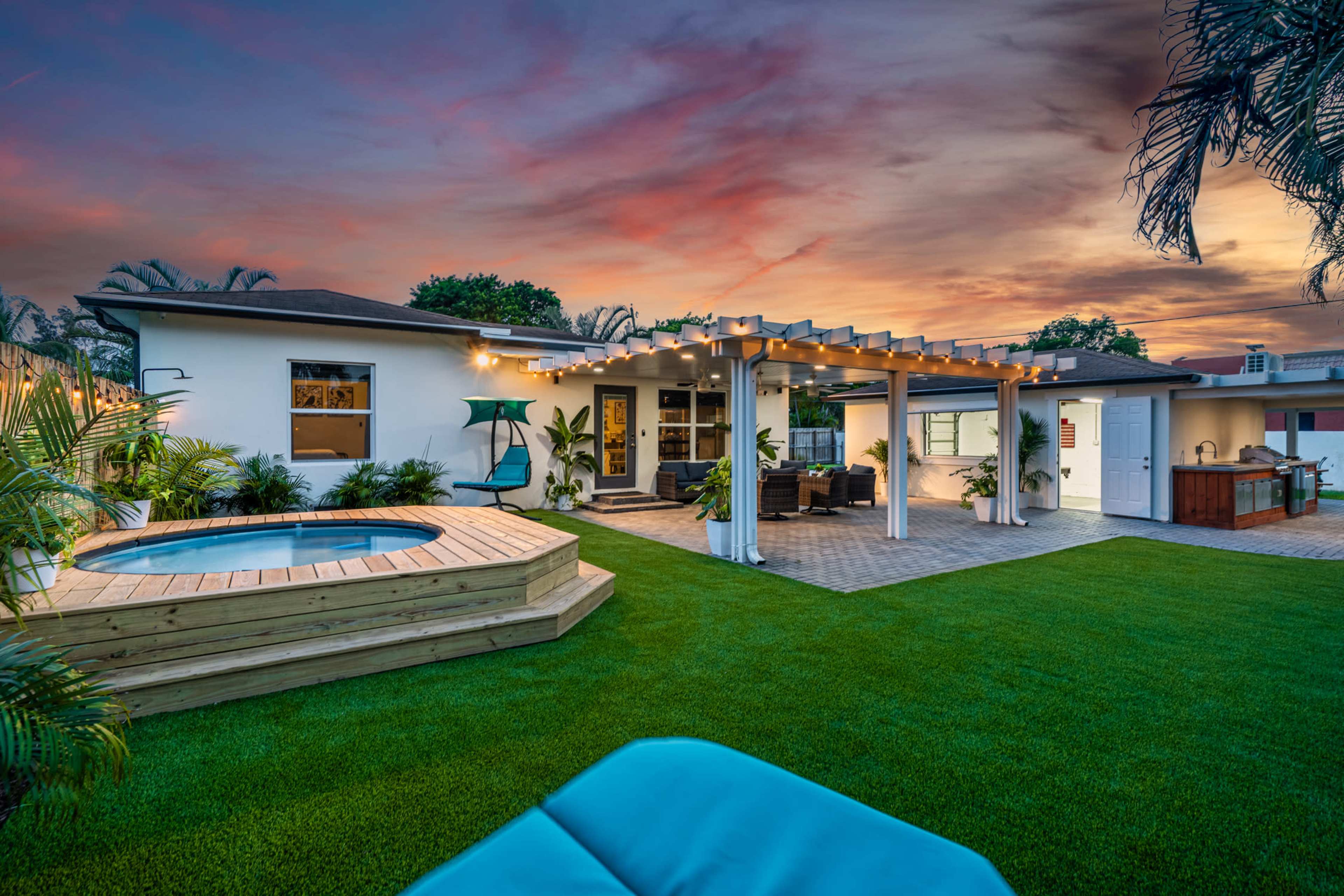 A backyard with a wooden deck, a hot tub, and a spacious lawn, framed by a house with a pergola and patio furniture under a colorful sunset sky.