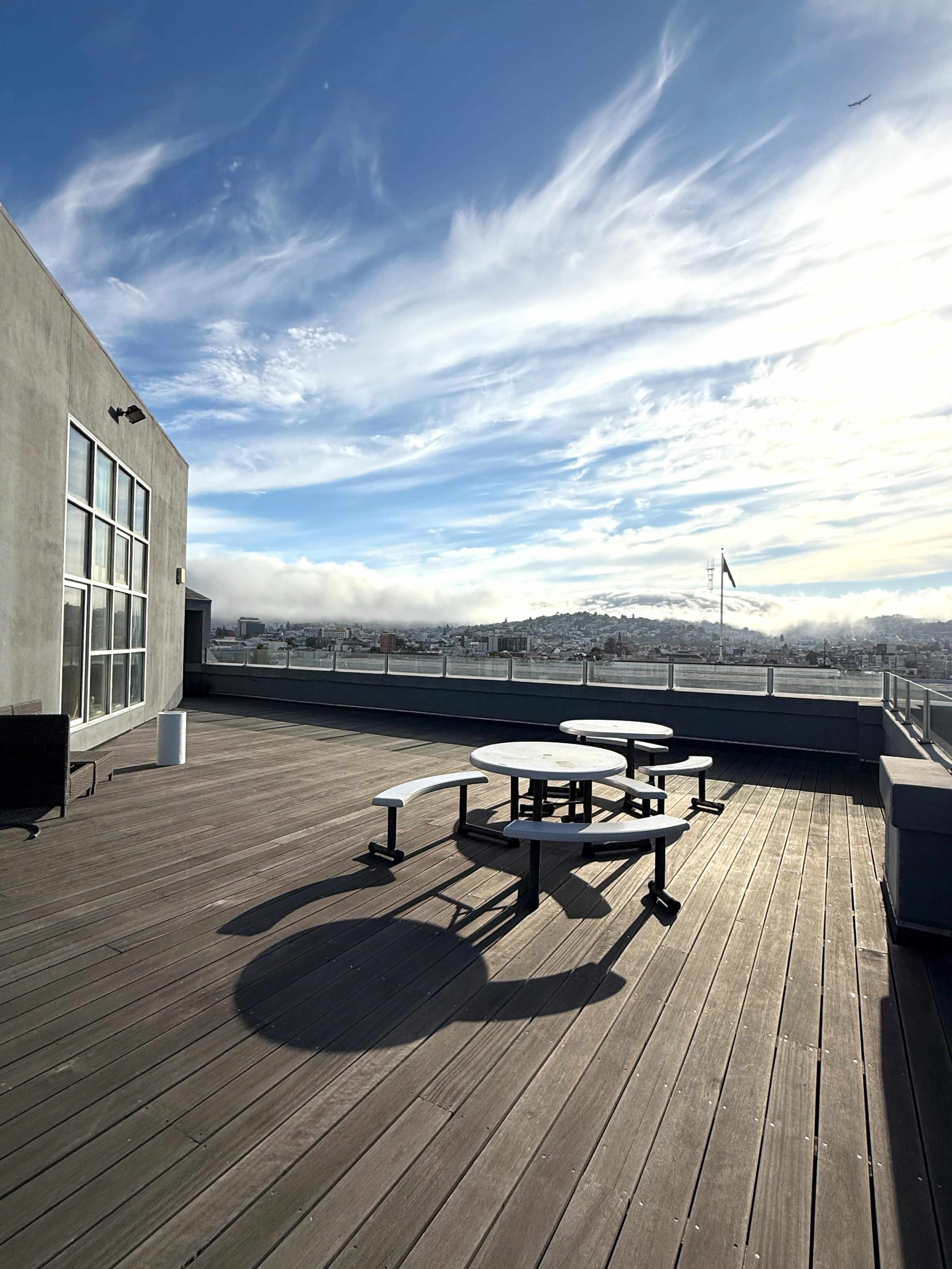 A wooden rooftop deck features a circular table and benches, with a view of the skyline and clouds in the background.
